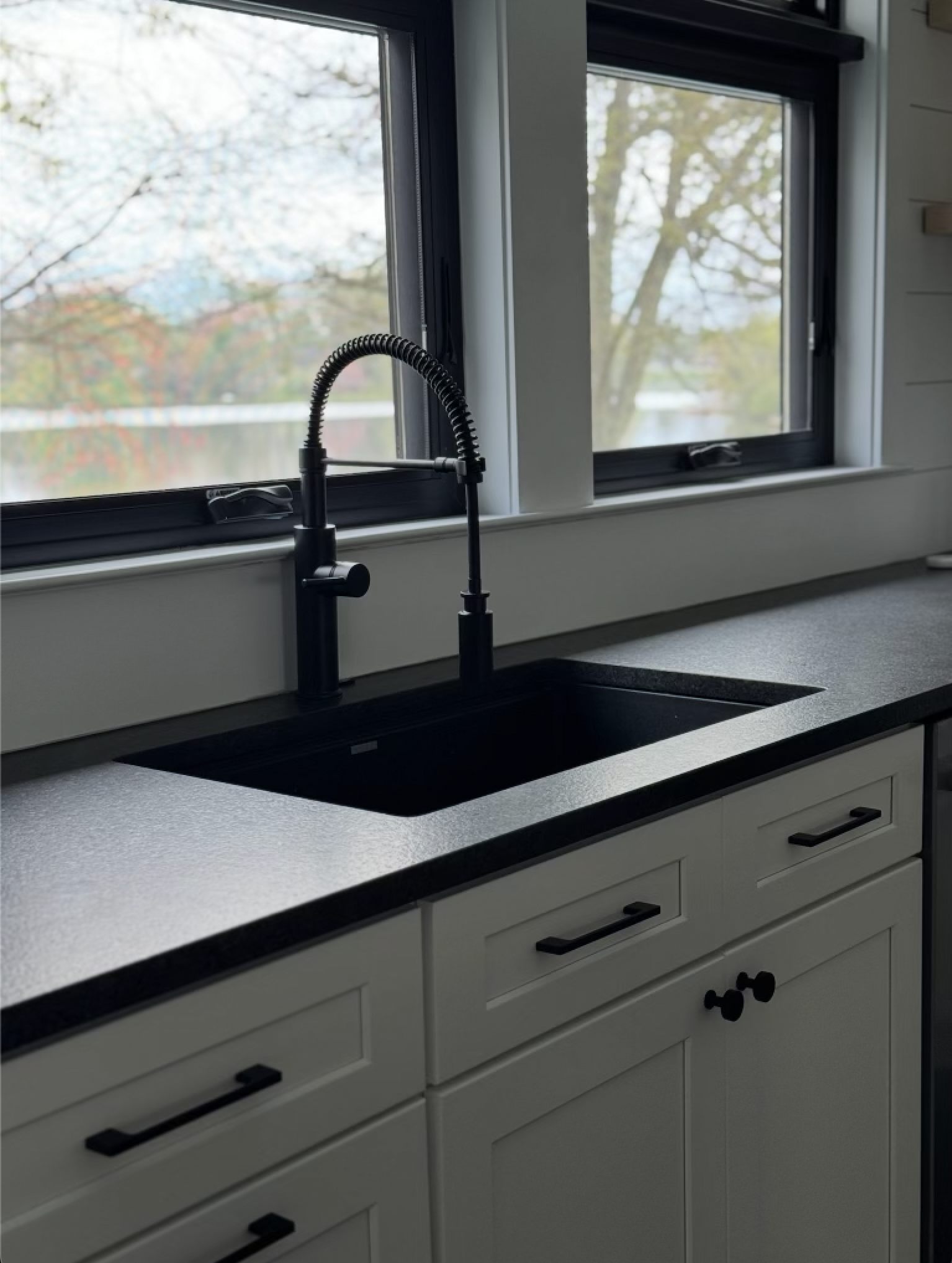 Kitchen sink with black faucet, white cabinetry, and large window showing trees outside.