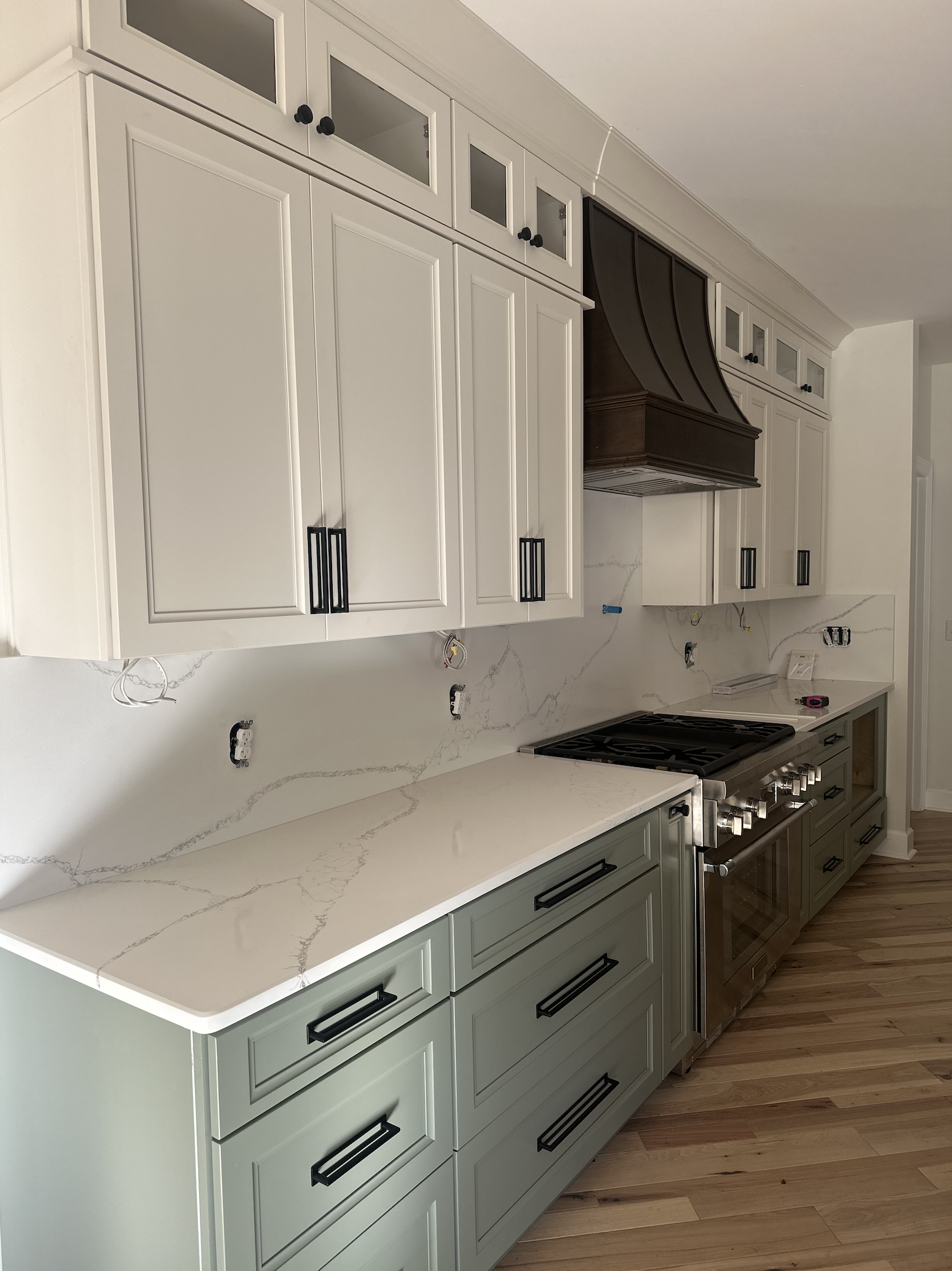 Kitchen with white upper cabinets, gray lower cabinets, a marble countertop, and a stainless steel stove. There is a dark brown range hood above the stove and electrical outlets on the wall.