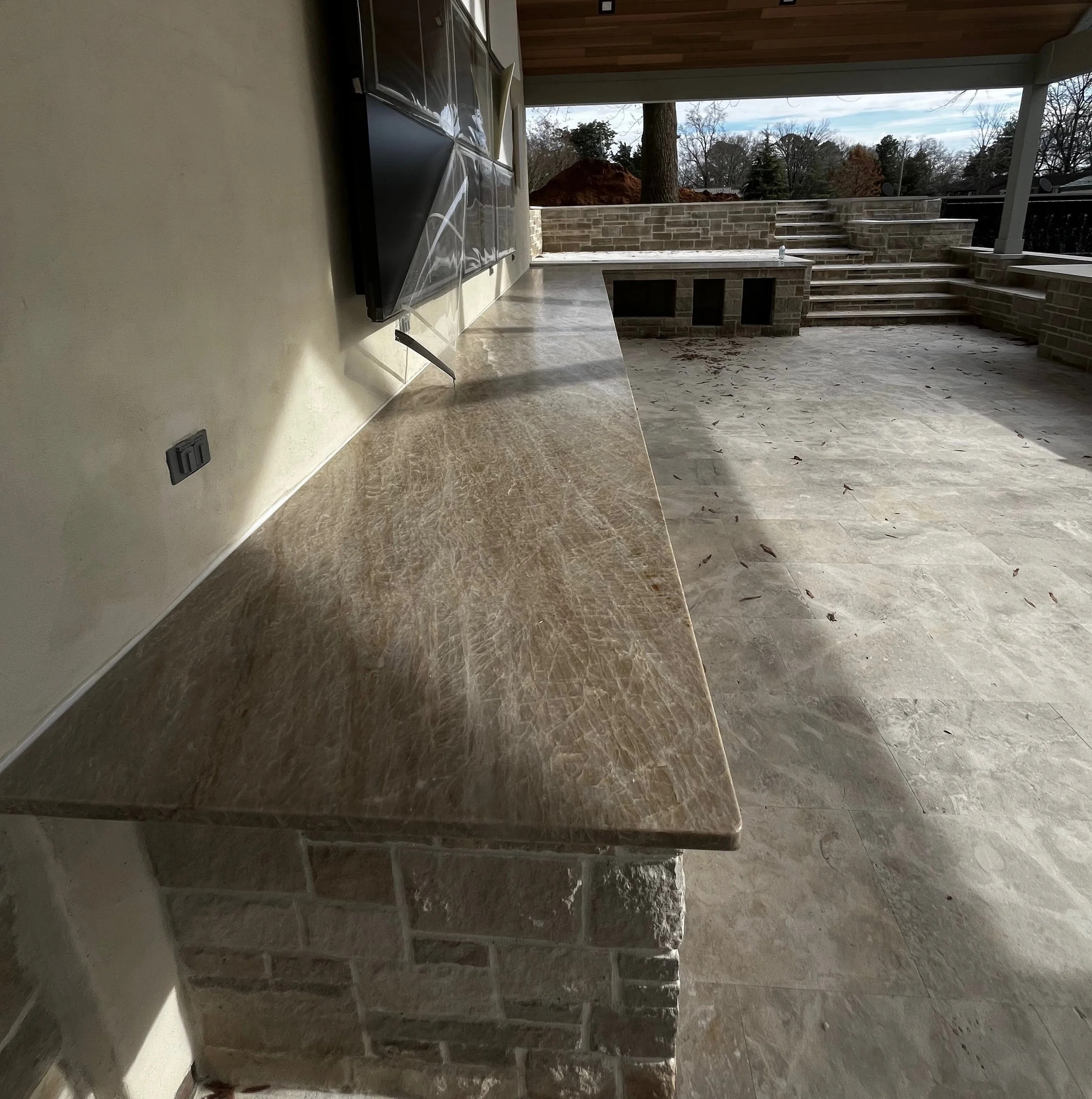Outdoor patio area with a long stone countertop, brick base, and steps leading to a lower level with a view of trees and a cloudy sky.