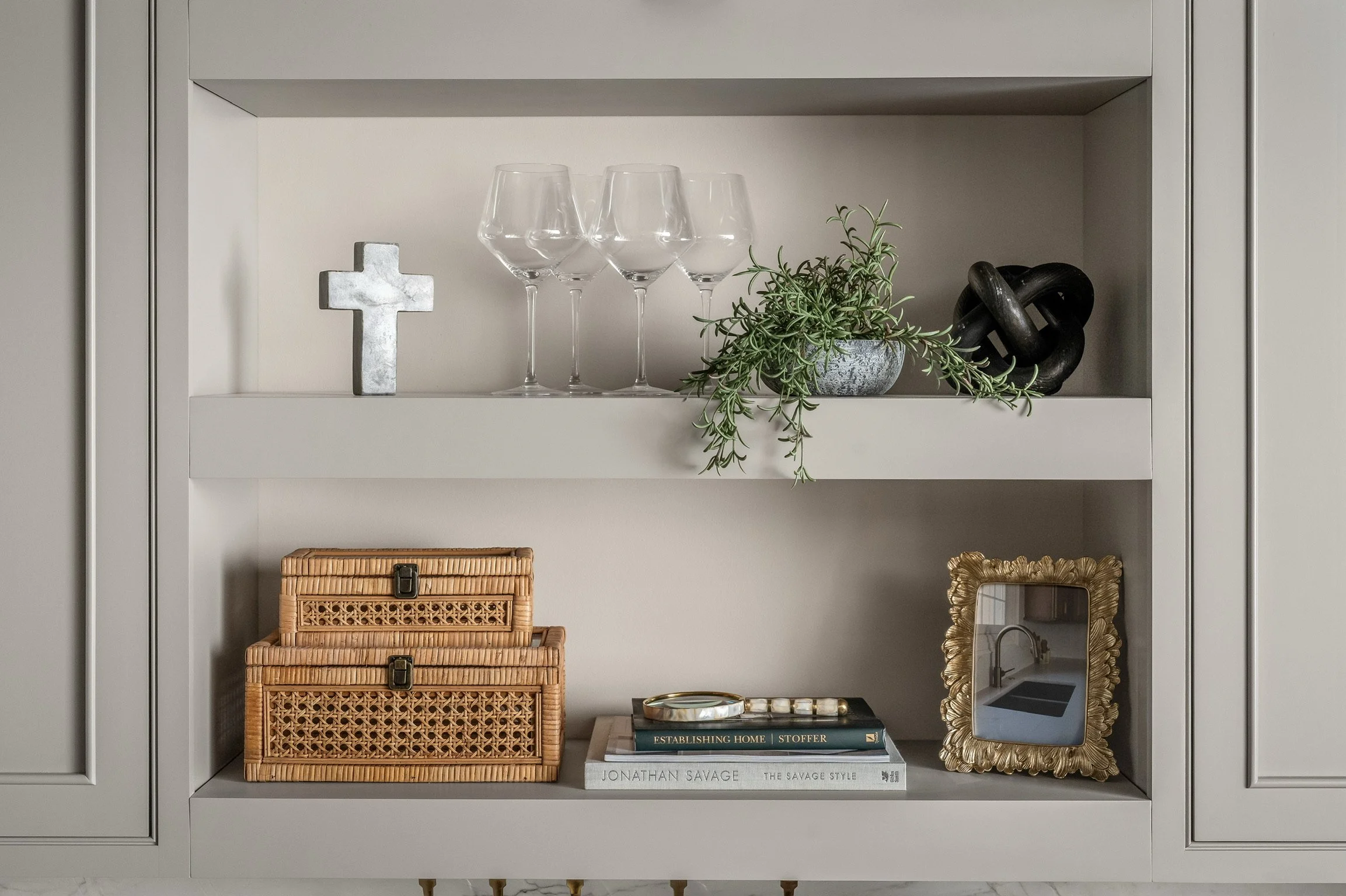 Open shelving with decorative items including a cross, four wine glasses, a potted plant, a black sculptural knot, two woven baskets, a stack of books, and a gold-framed mirror.