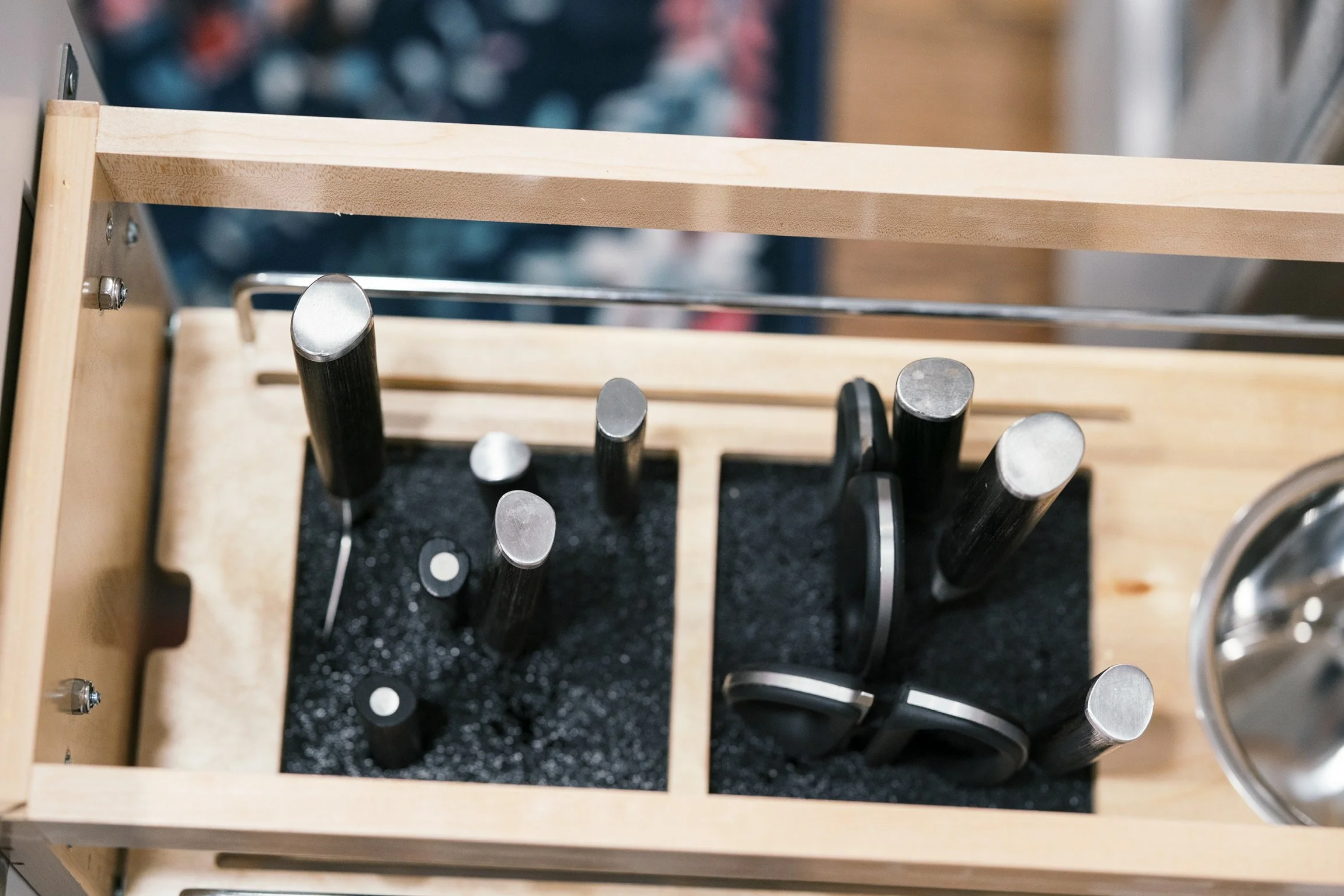 A wooden knife block with black-handled kitchen knives and scissors, placed on a black foam mat inside a wooden holder.