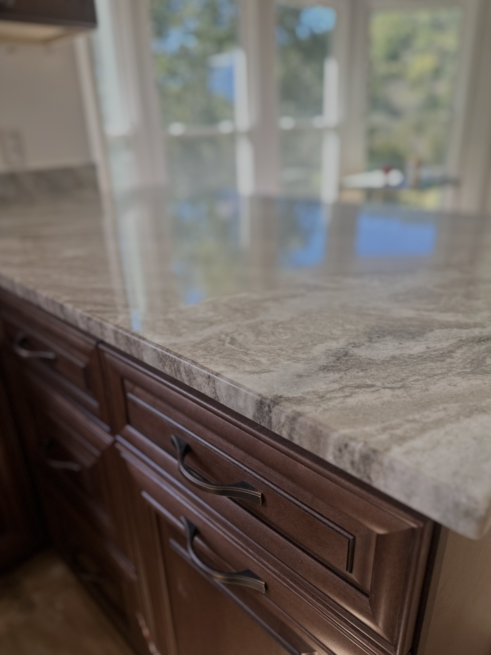 Close-up of a kitchen countertop made of beige and gray patterned stone, with dark wood cabinets underneath and a blurred background of large window with trees outside.
