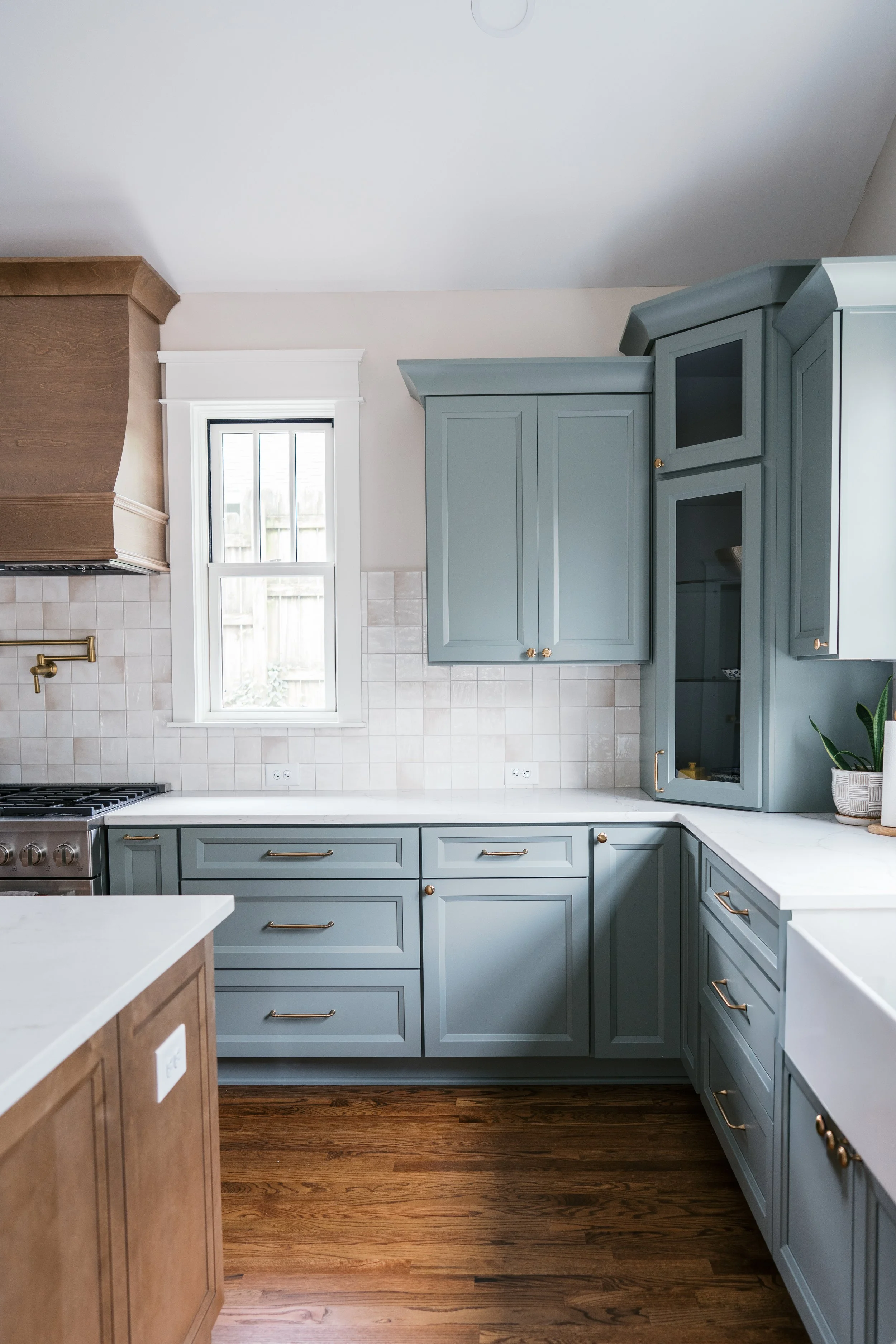 A kitchen with light blue cabinets, a white marble countertop, a small window, and hardwood floors.