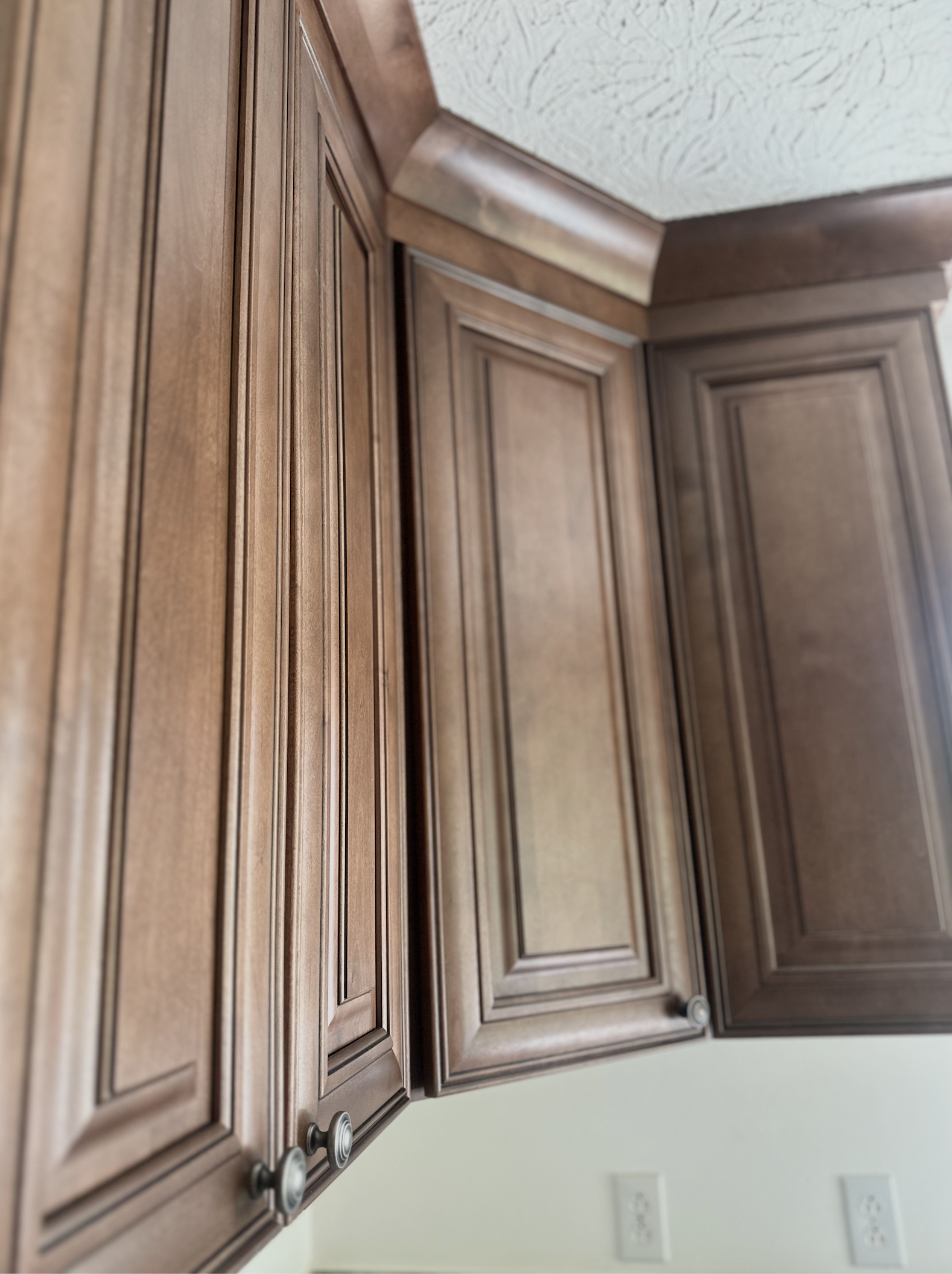 Close-up of wooden kitchen cabinets with decorative panels and round knobs, with electrical outlets visible on a light-colored wall.