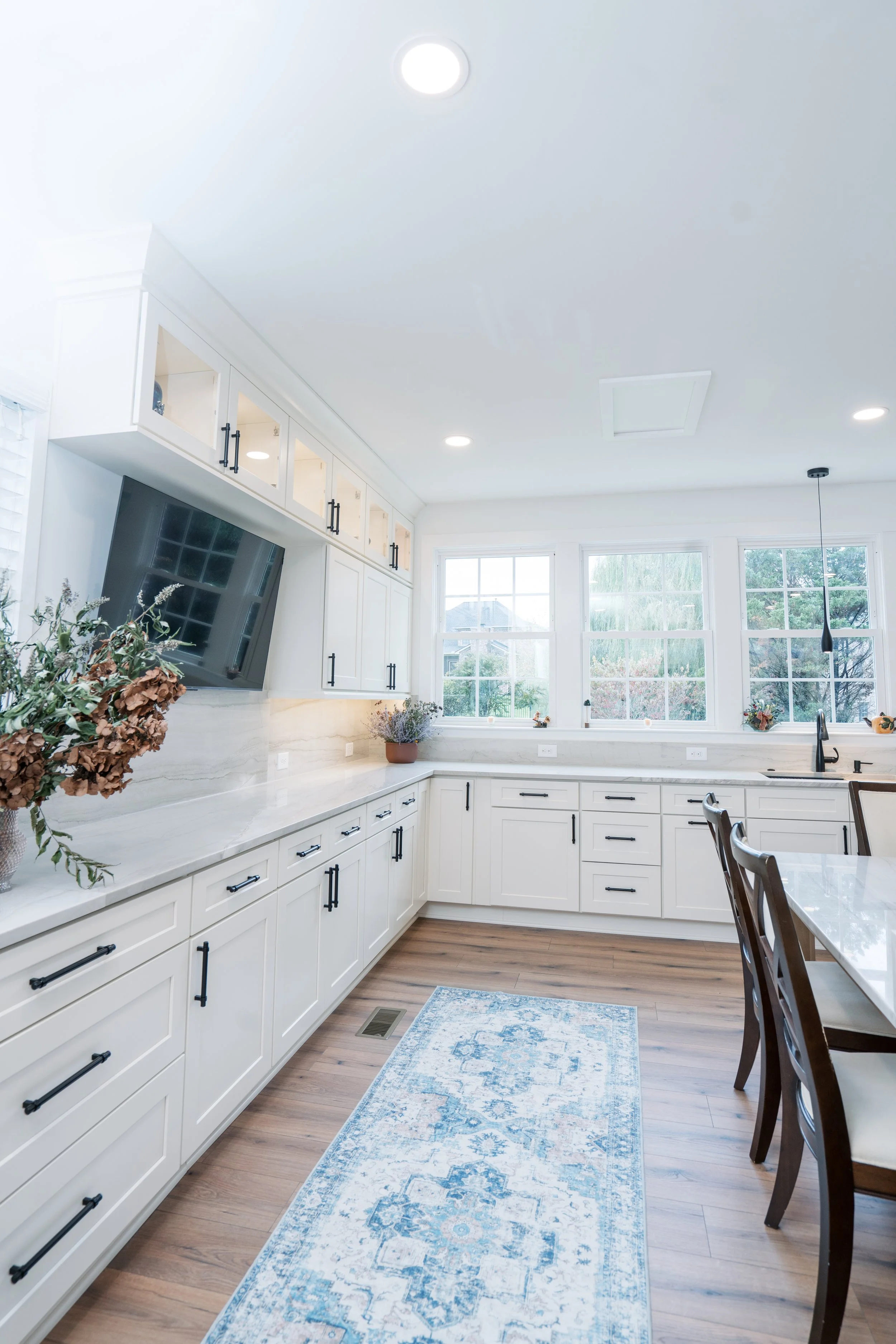 Bright kitchen with white cabinets, wooden floor, large windows, a TV mounted on the wall, and a blue-patterned rug on the floor.