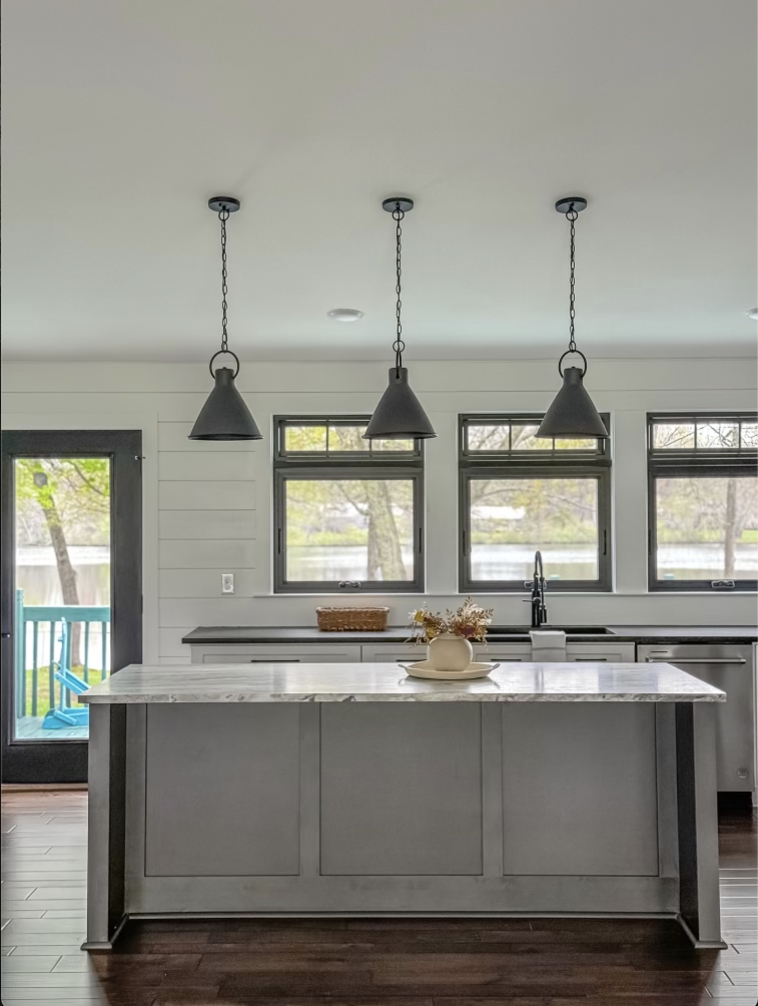 Modern kitchen with pendant lighting, a marble countertop island, and three windows overlooking trees and a yard.