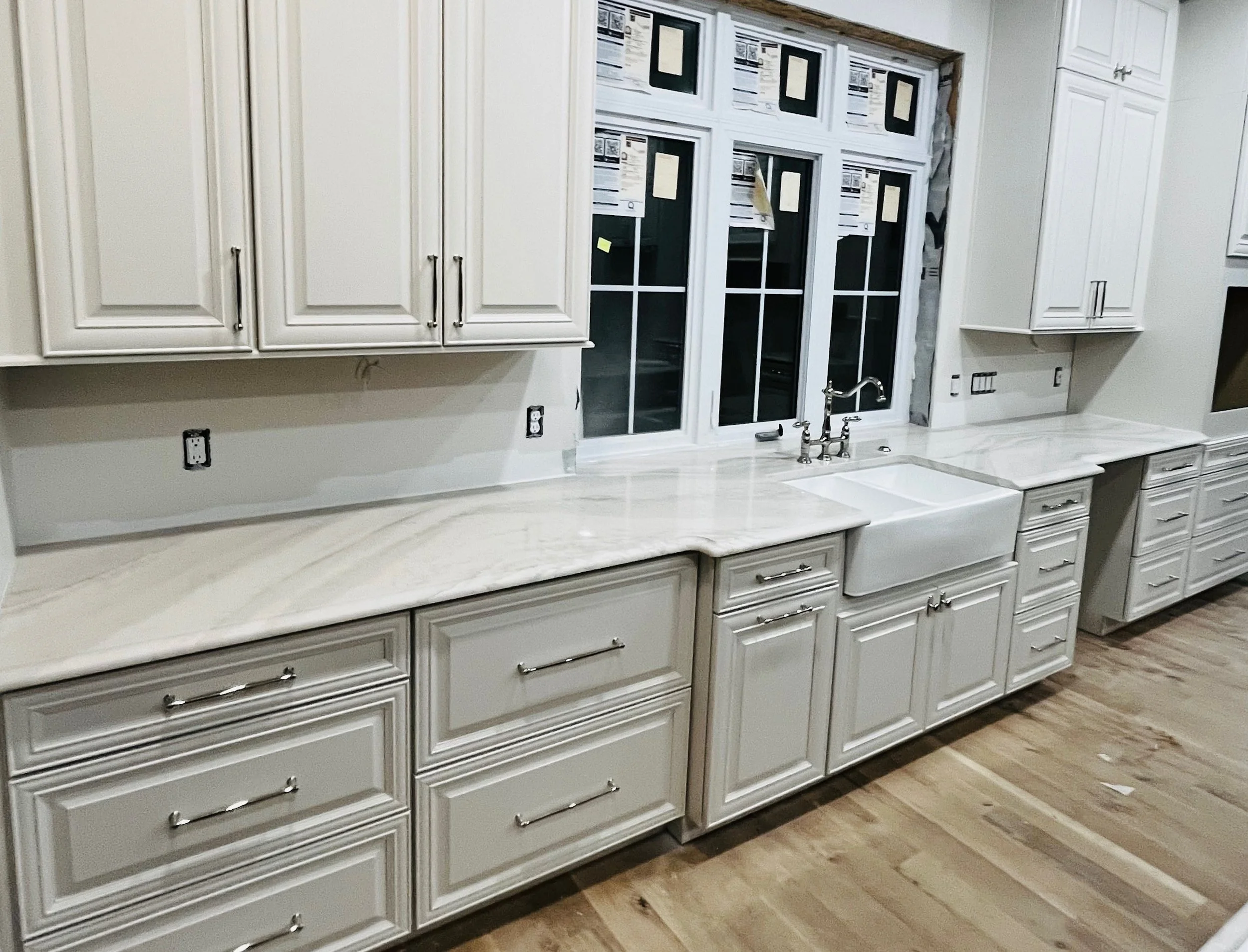 Kitchen with white cabinetry, marble countertops, and a farmhouse sink under a window. The kitchen appears to be under construction or renovation, with open electrical outlets and unfinished areas around the window.