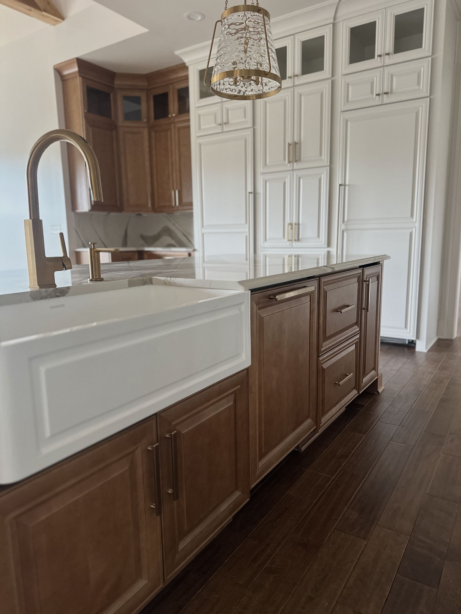 Kitchen with brown and white cabinetry, marble countertops, a gold and glass pendant light, and a farmhouse sink with a gold faucet.