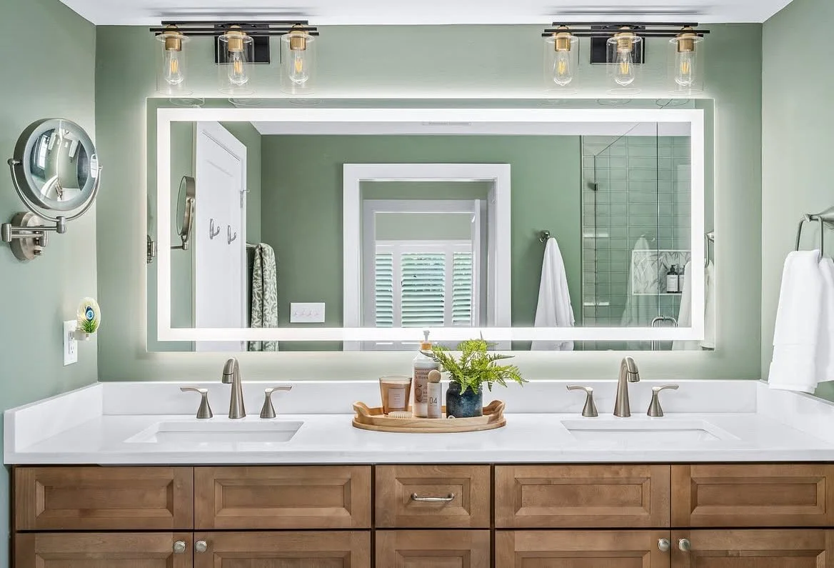 Bathroom with a large illuminated mirror above a double sink vanity, wooden cabinet, green walls, and decorative items on a tray