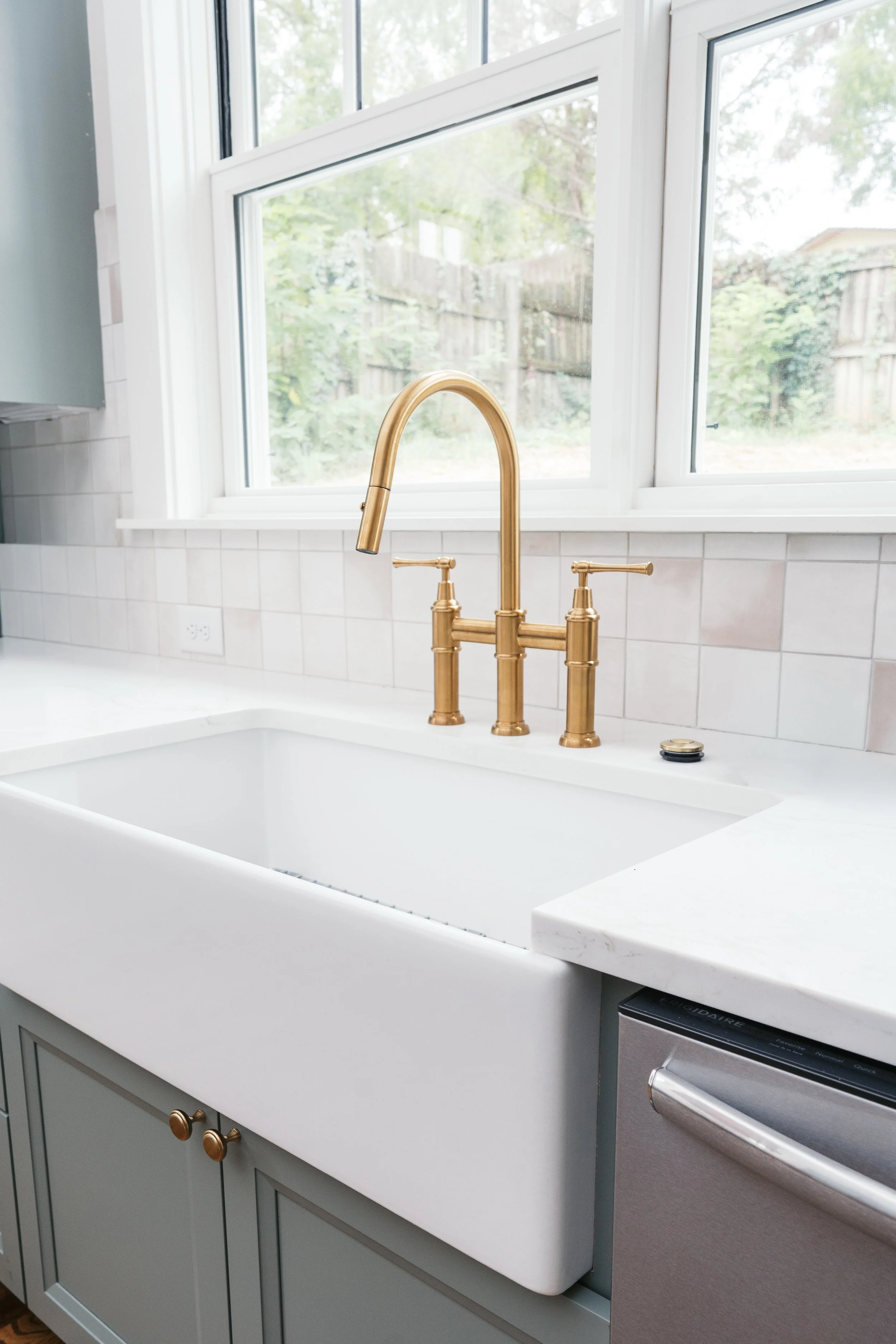 Kitchen sink with a white farmhouse basin, gold faucet, and a window showing greenery outside.