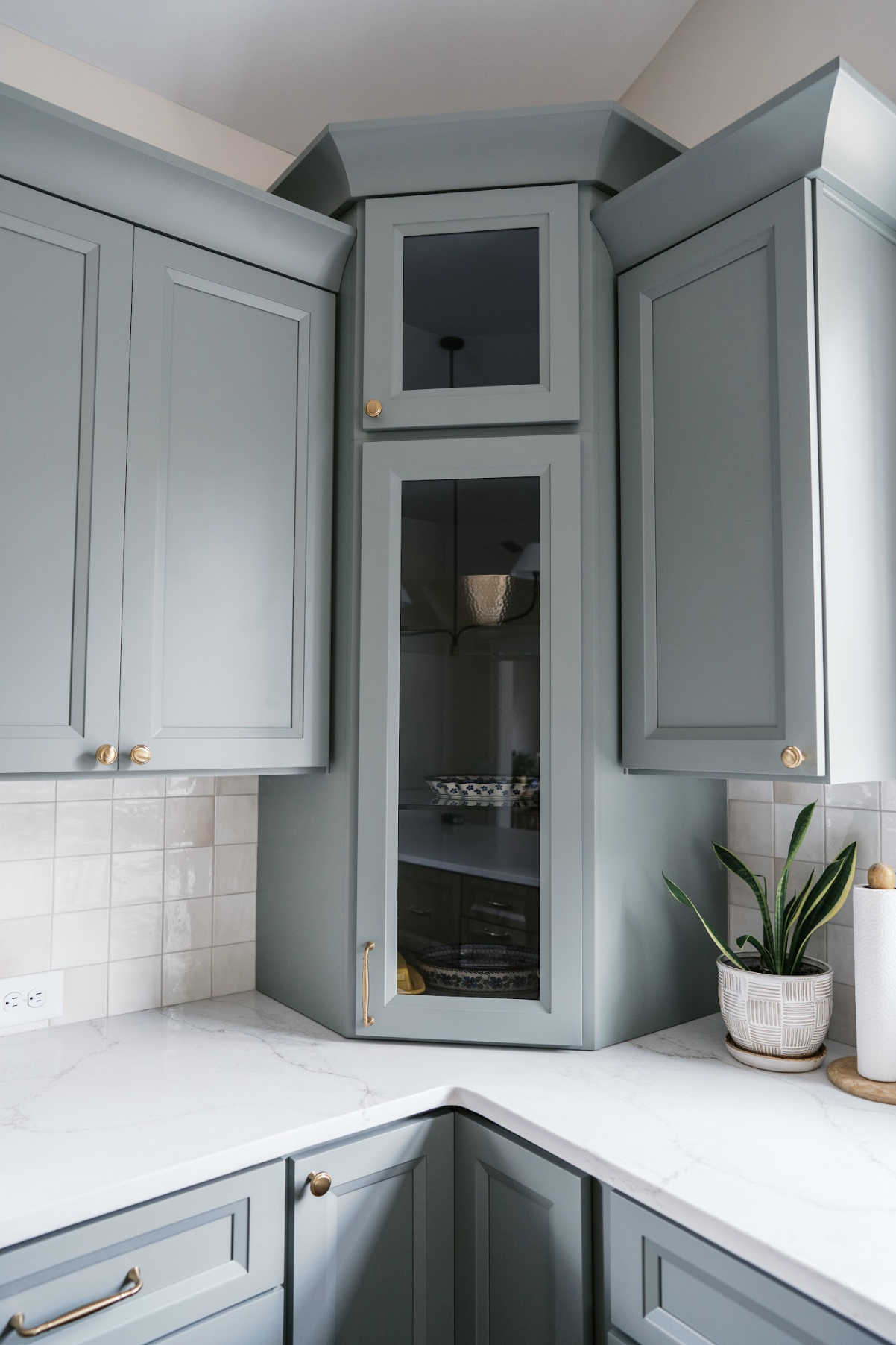 Close-up of light blue kitchen cabinets with gold knobs, marble countertop, and a potted plant next to a paper towel roll.