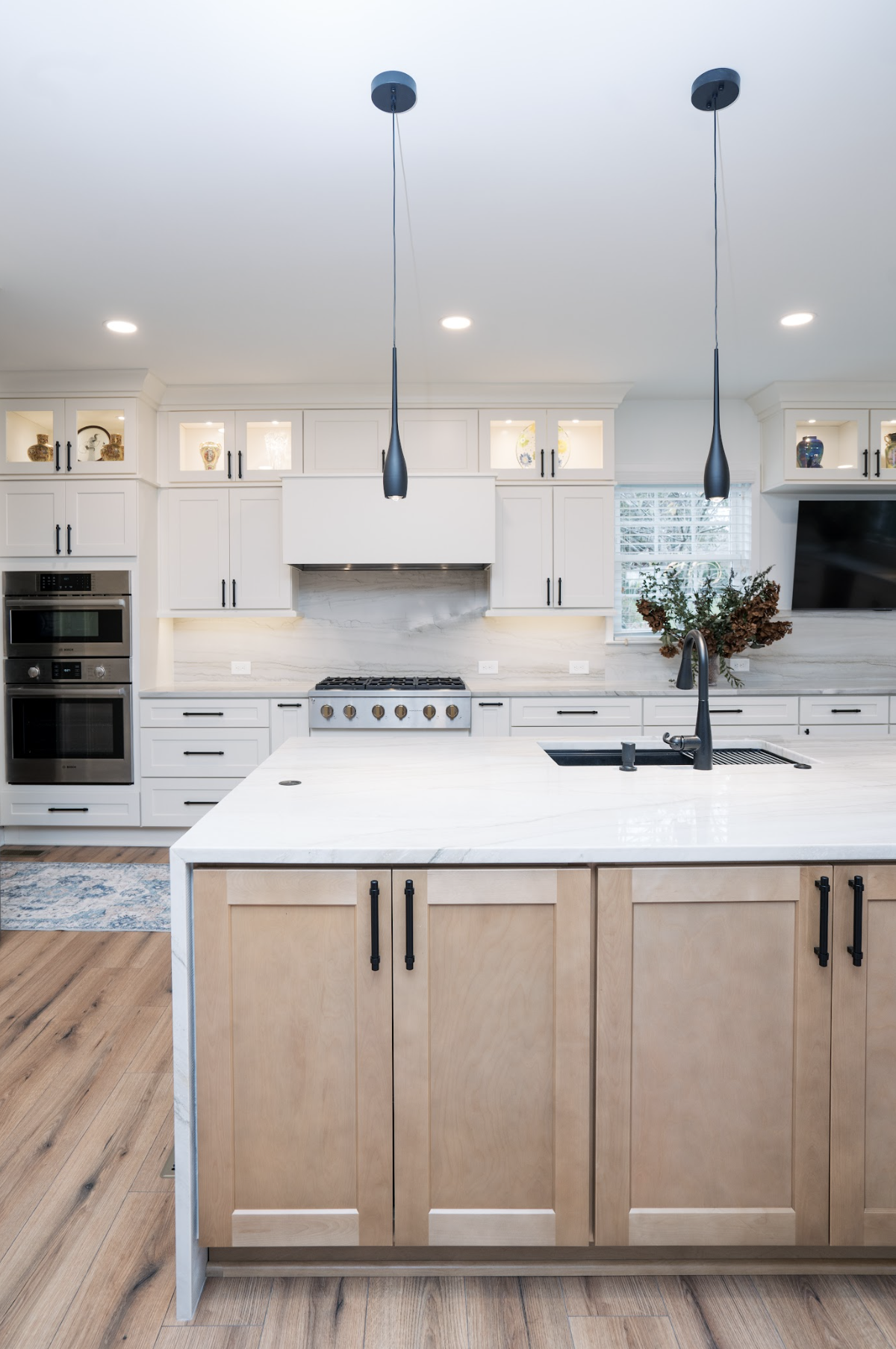 Modern kitchen with white cabinets, a central island with a light wood finish, black fixtures, a marble backsplash, built-in ovens, a stove, pendant lighting, and a wall-mounted television.