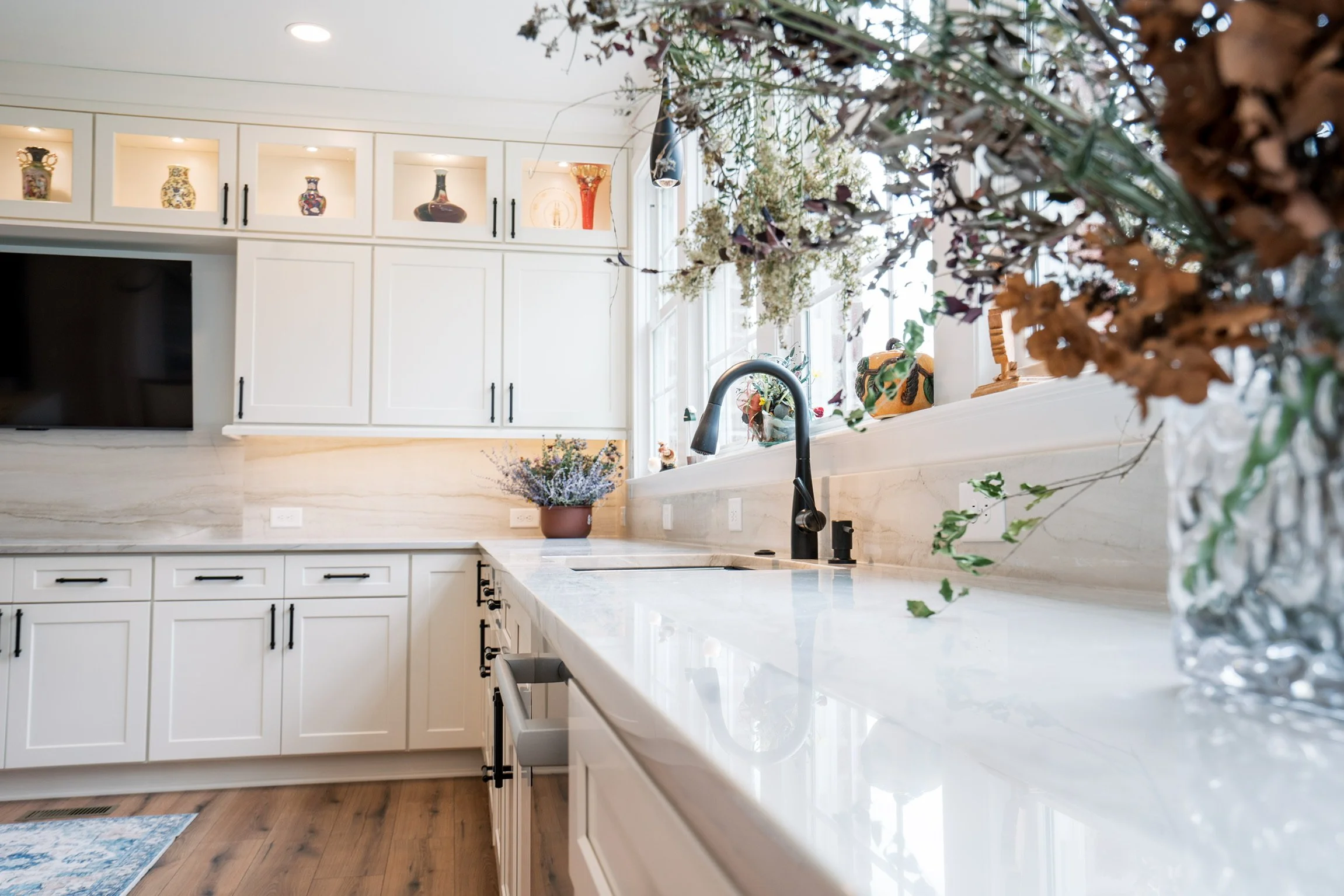 A bright kitchen with white cabinets, a marble countertop, black hardware, a black faucet, and floral arrangements on the counter and window sill.