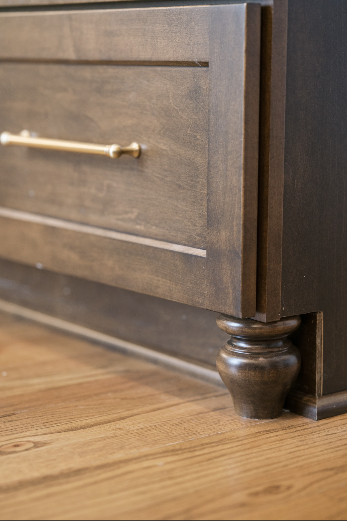 Close-up of a wooden dresser with a bronze-colored knob and turned wooden leg on a hardwood floor.