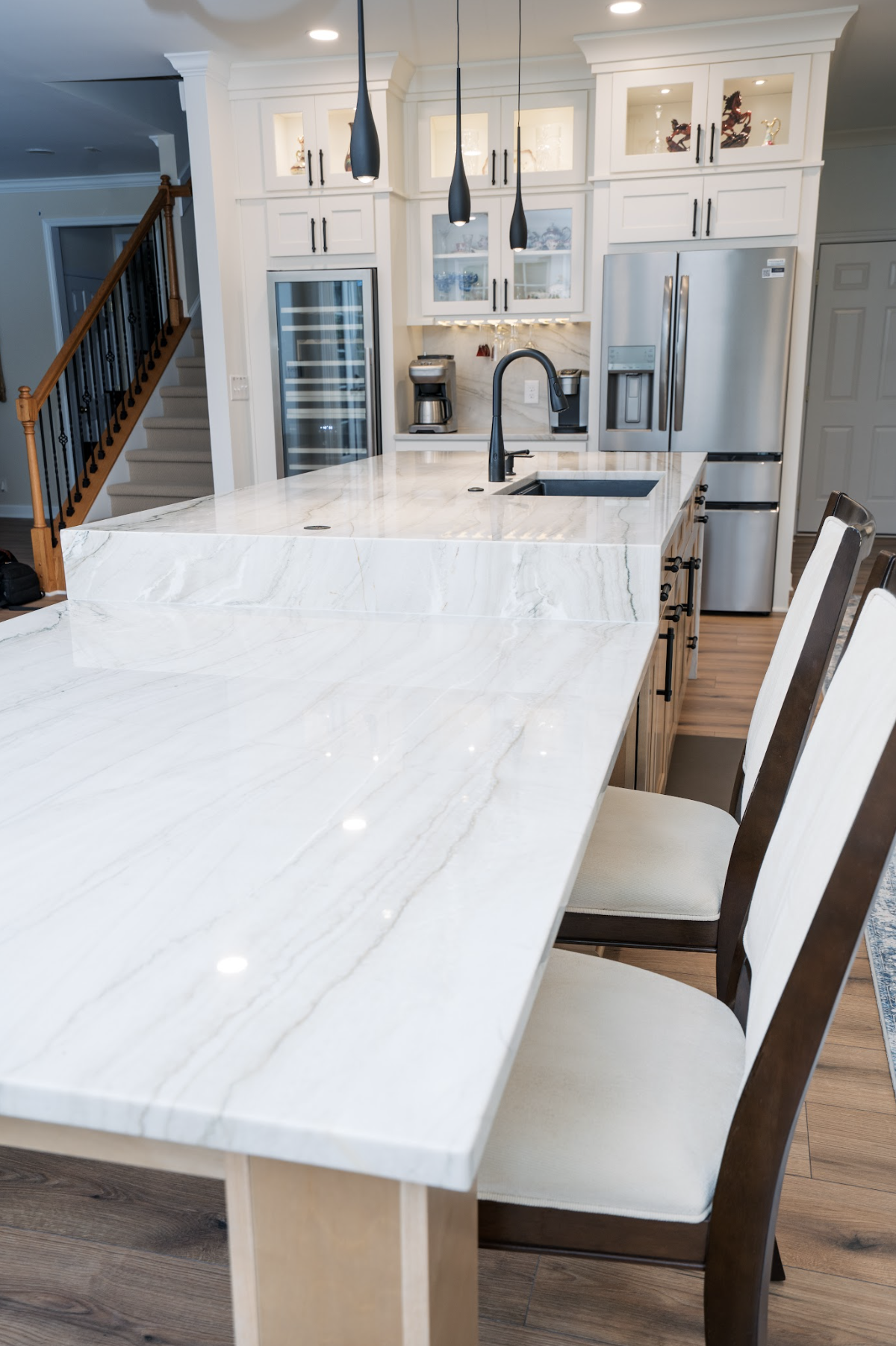 Modern kitchen with a white marble countertop, black sink faucet, white cabinetry with black handles, stainless steel refrigerator, wine cooler, coffee maker, and pendant lights.