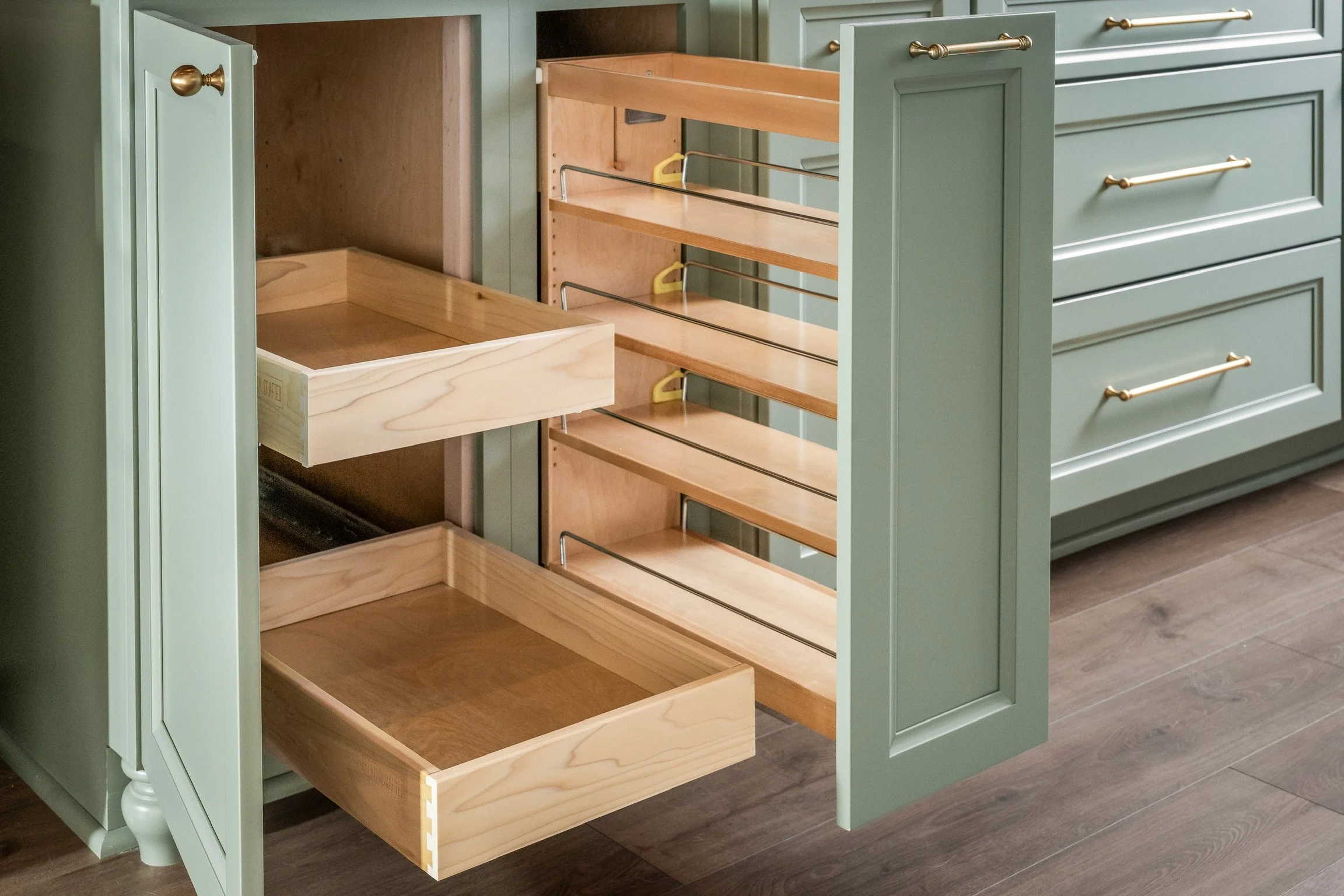 An open green kitchen cabinet with wooden drawers and shelves inside, set on wooden flooring.