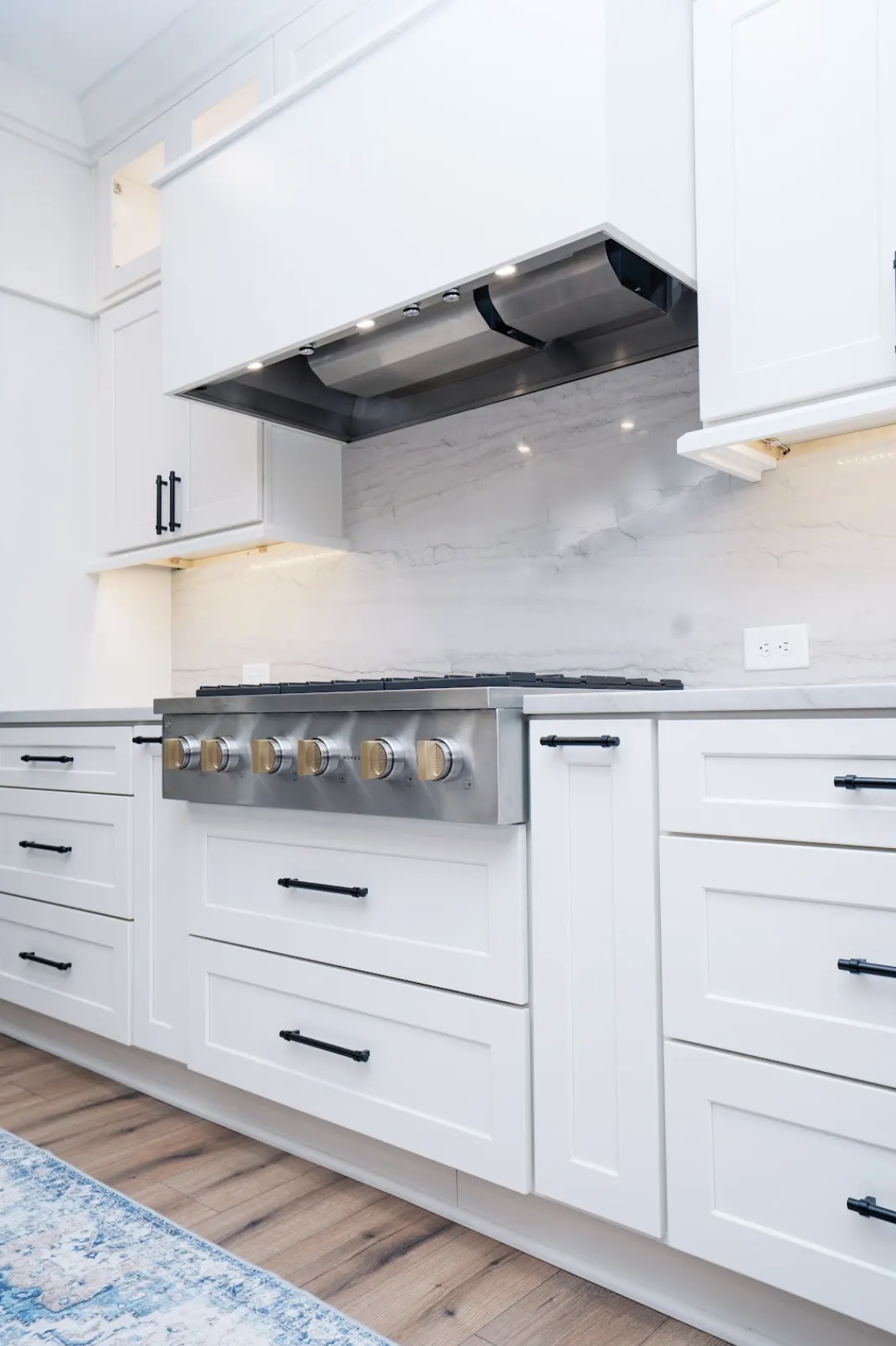 Modern kitchen with white cabinets, black handles, a stainless steel stove with golden knobs, a stainless steel range hood, and a white marble backsplash.