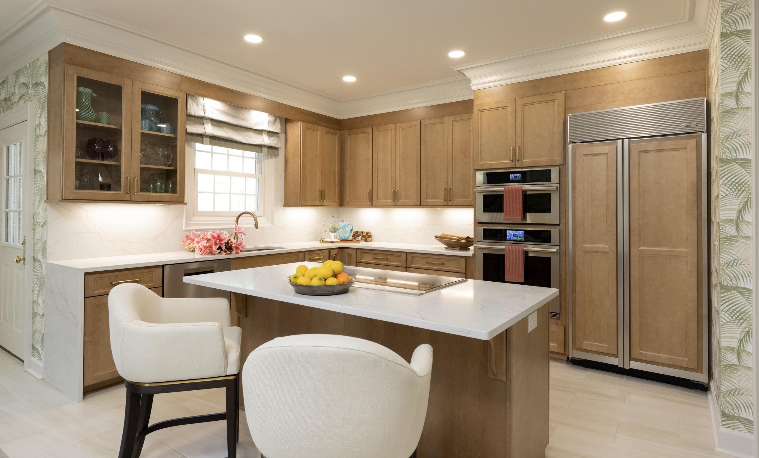 Modern kitchen with wooden cabinets, white marble countertops, a central island with a bowl of lemons, and built-in double ovens; a window above the sink with beige Roman shades and decorative pink flowers on the countertop.