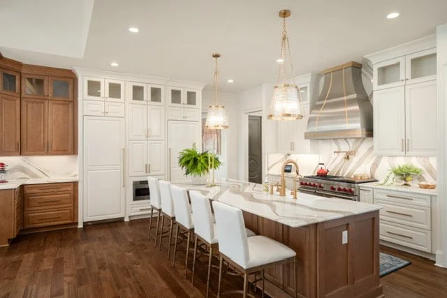 Modern kitchen with white cabinets, a large marble island, and wooden accents. Four white chairs sit at the island, with pendant lights hanging above. Stainless steel range with a hood, a plant, and decorative items are on the countertops.