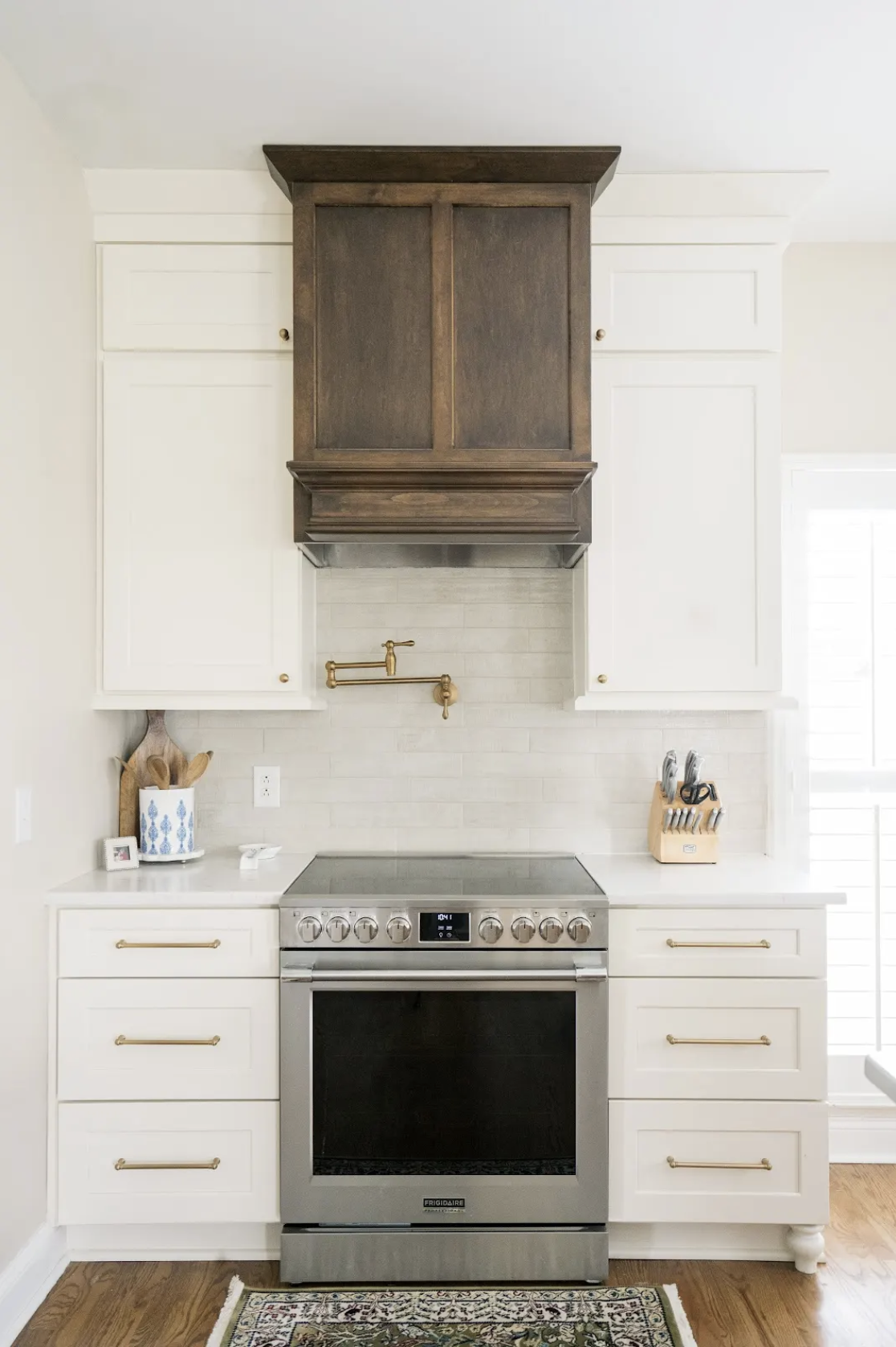 Kitchen with white cabinets, gold handles, stainless steel oven, wooden cutting board, blue and white utensil holder, and knife block, with a wooden range hood and a beige tiled backsplash.