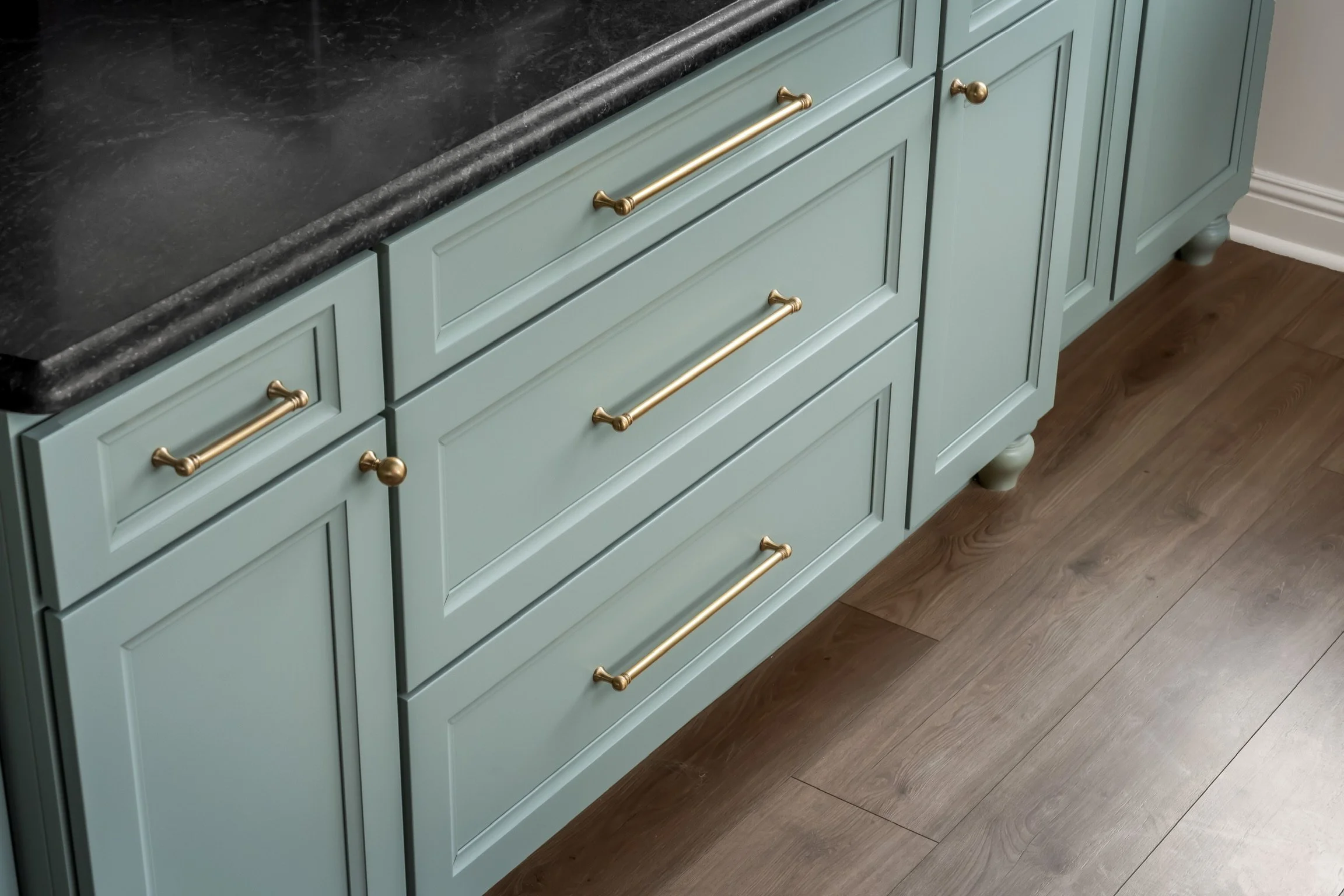 Light blue kitchen cabinets with gold handles and a dark countertop, with wood flooring.