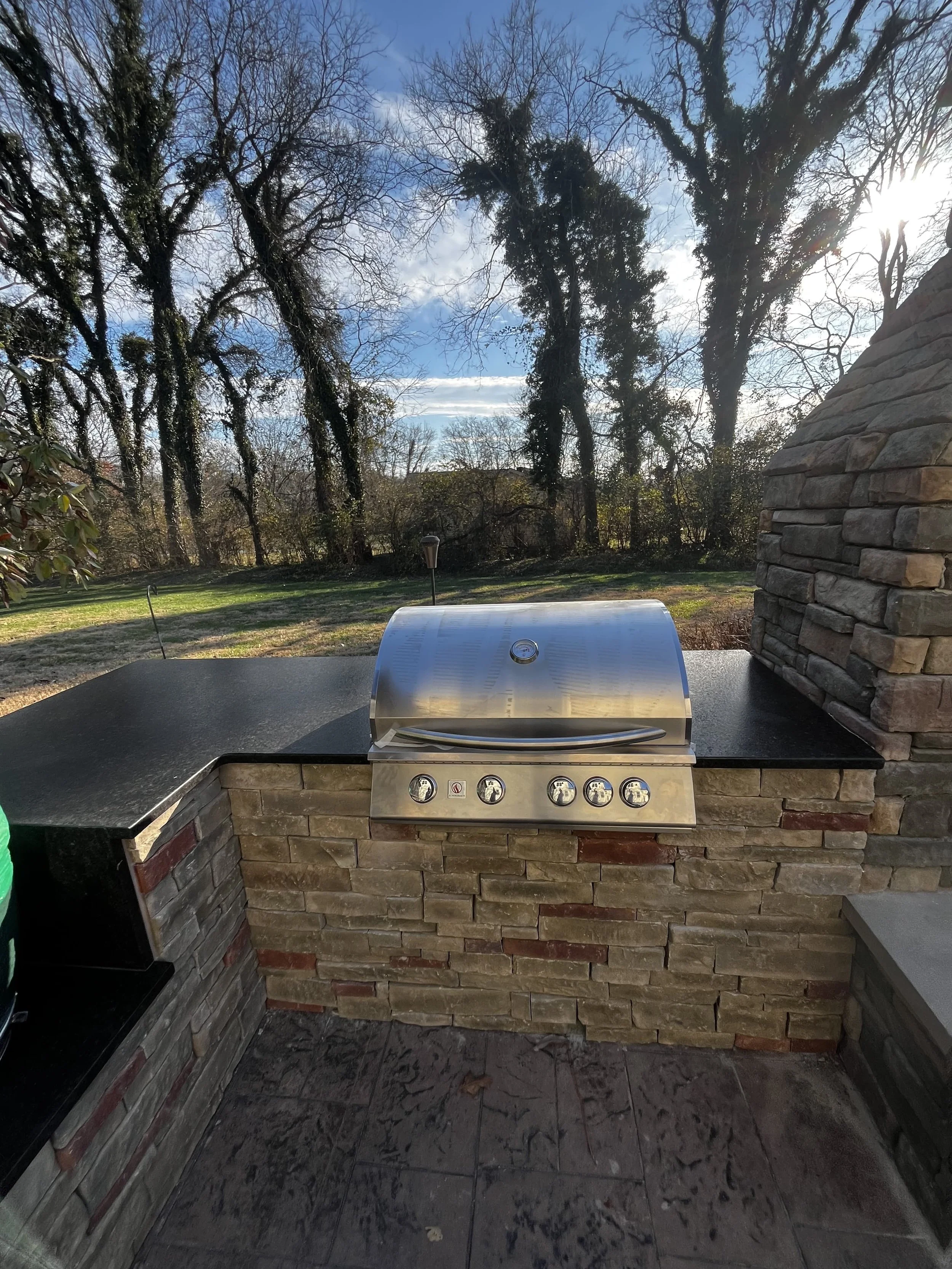 Outdoor stainless steel grill on a brick and stone fire pit, with a black countertop and stone chimney, overlooking a yard with trees and a partly cloudy sky.