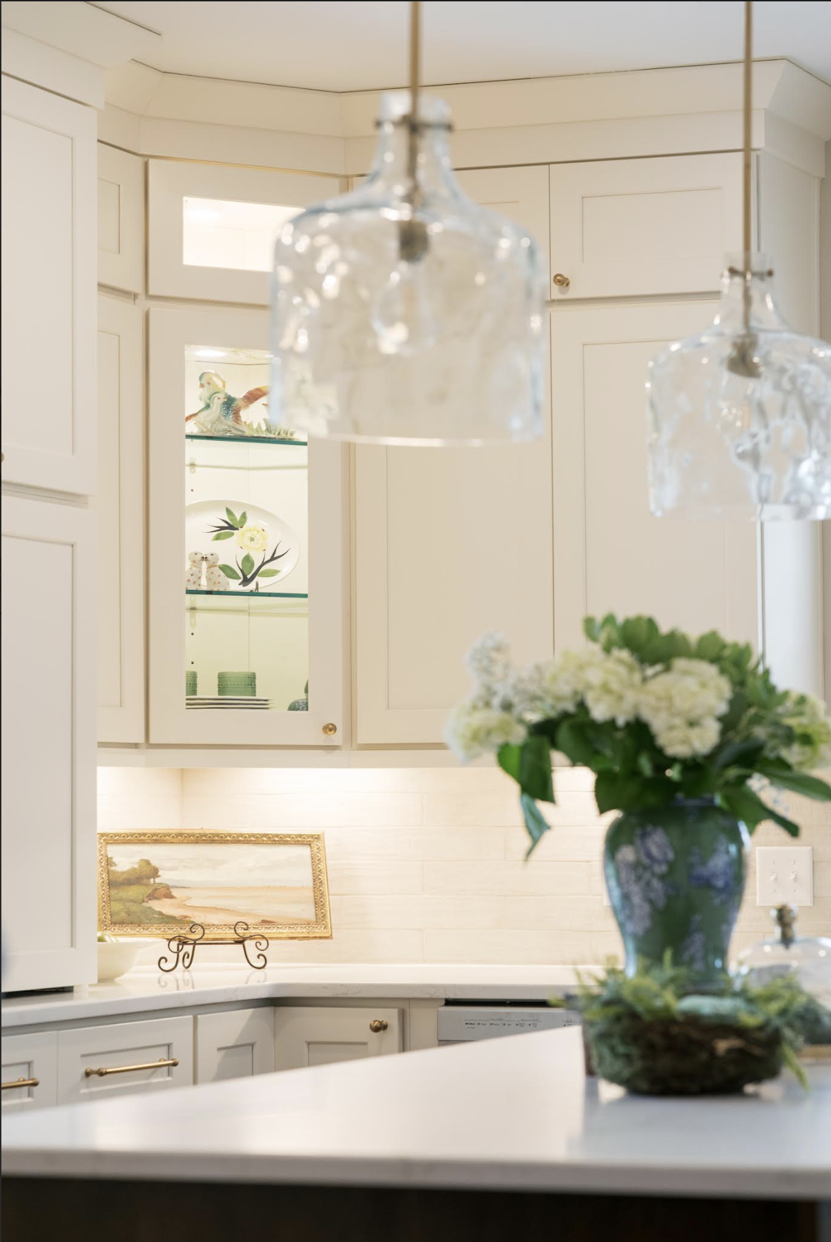 A kitchen interior featuring white cabinets, a marble countertop, hanging glass pendant lights, a floral arrangement in a blue vase on the counter, and decorative items on a glass shelf and the wall.