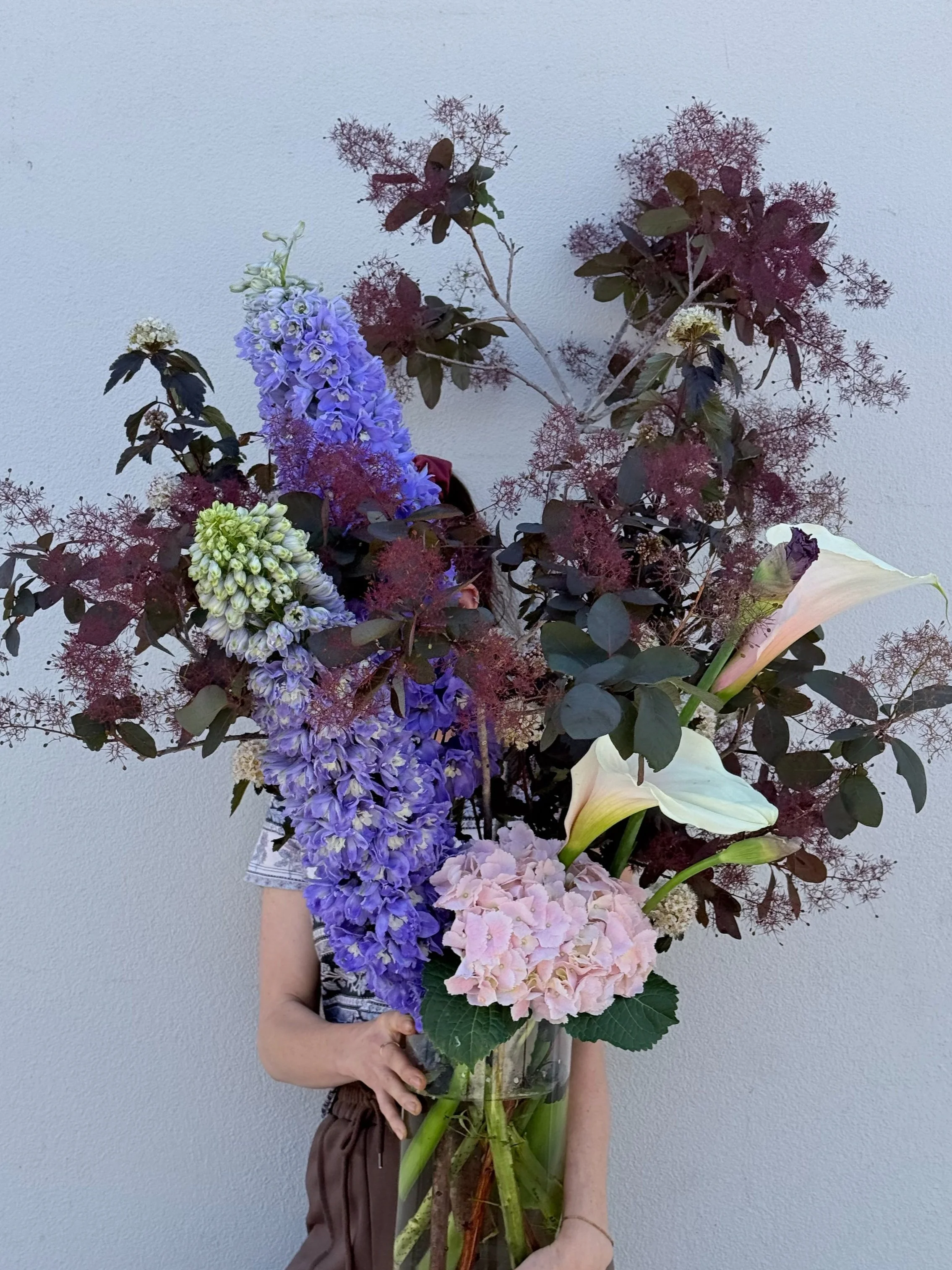 A person holding a large bouquet with purple delphiniums, pink hydrangeas, calla lilies, and dark foliage against a light gray background.