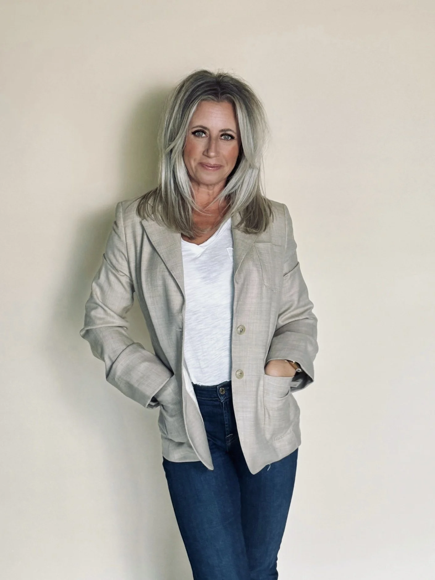 A woman with shoulder-length, layered blonde hair standing against a plain beige wall, wearing a light-colored blazer, white t-shirt, and dark jeans.