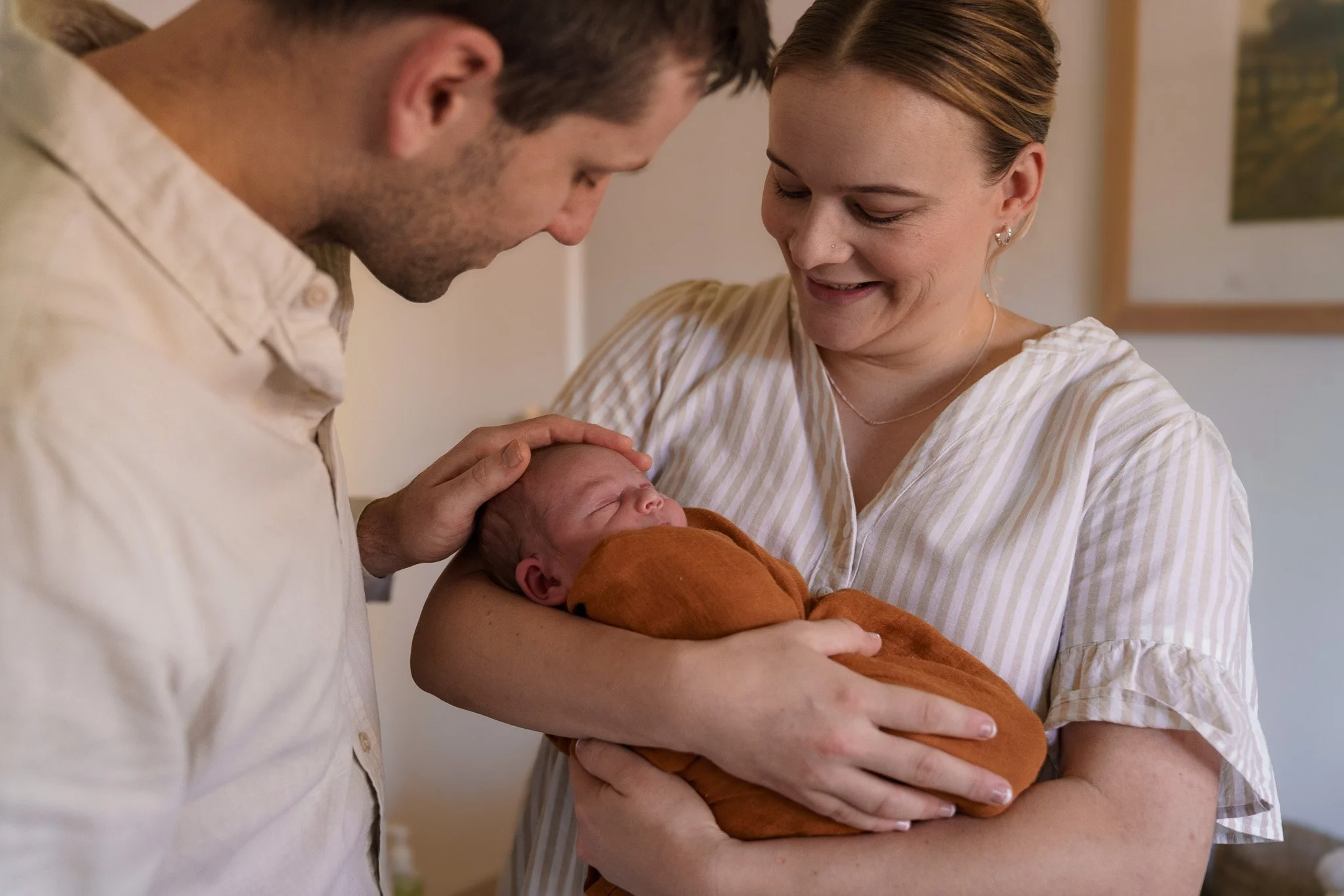 A man and a woman look lovingly at a sleeping newborn baby wrapped in an orange blanket, with the man gently touching the baby's head.