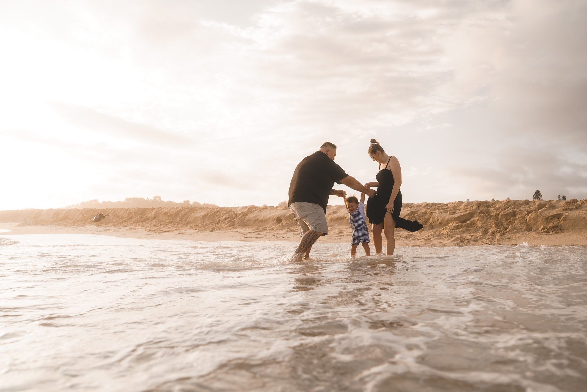 A family of three playing in shallow ocean water on a sandy beach during sunset.