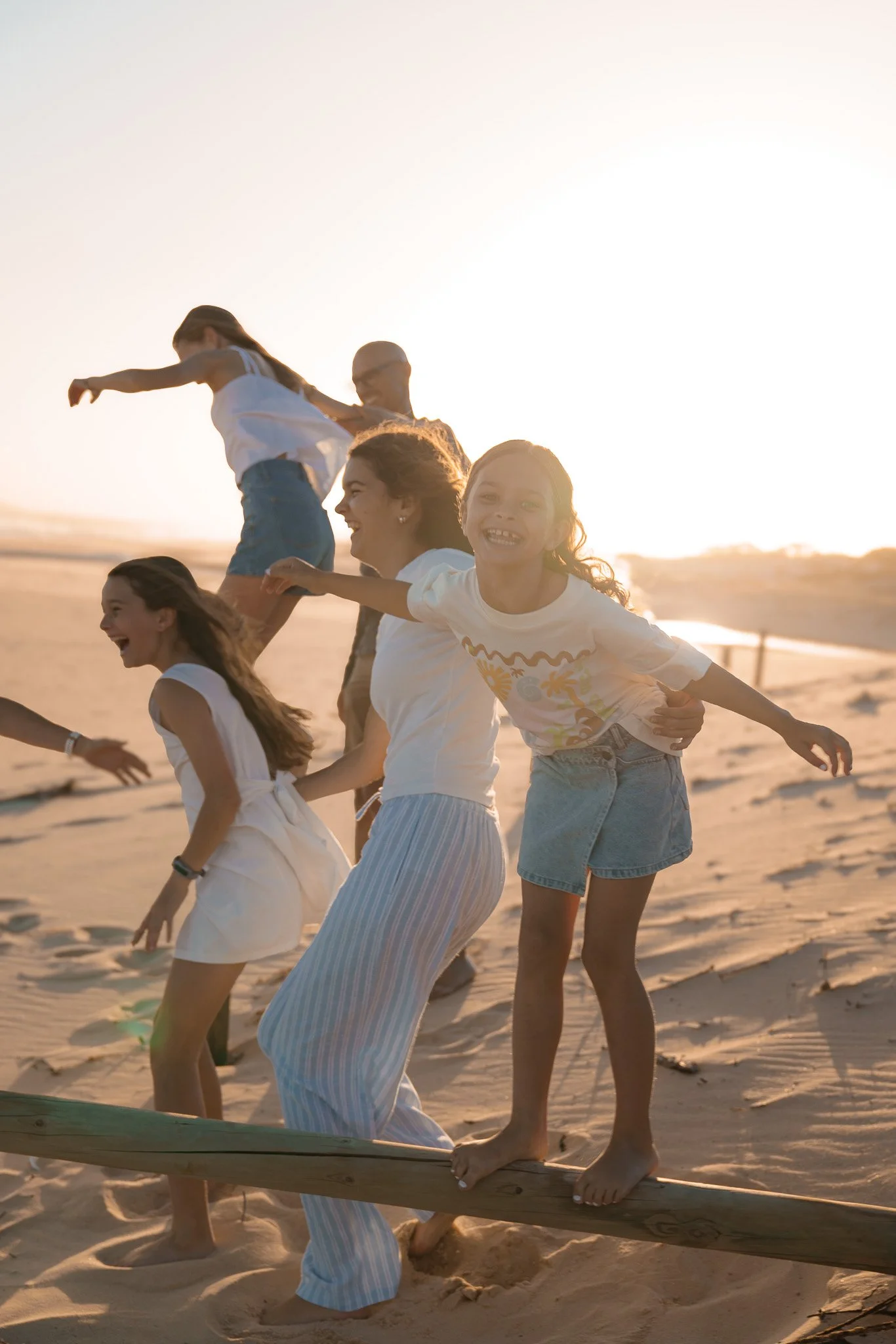 Group of children playing and balancing on a wooden beam at the beach during sunset.