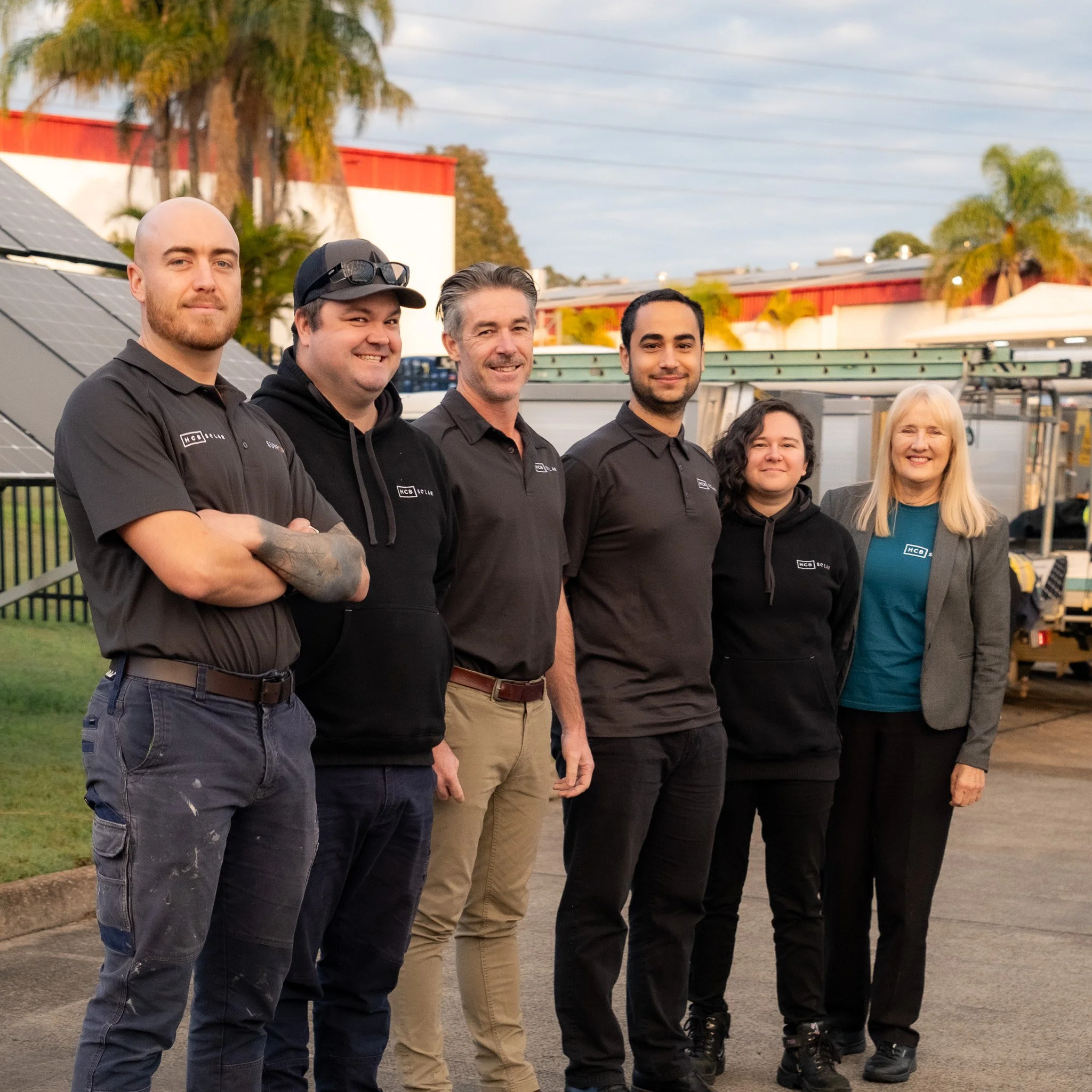 Group of six people standing outdoors, wearing black and teal clothing, with solar panel and utility vehicle in background.