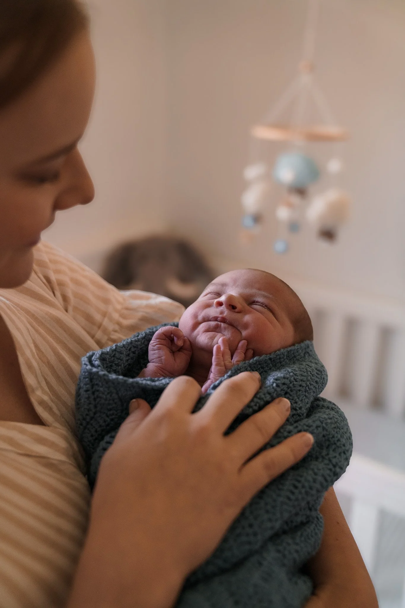 A woman holding a sleeping baby wrapped in a gray knitted blanket in a softly lit room.