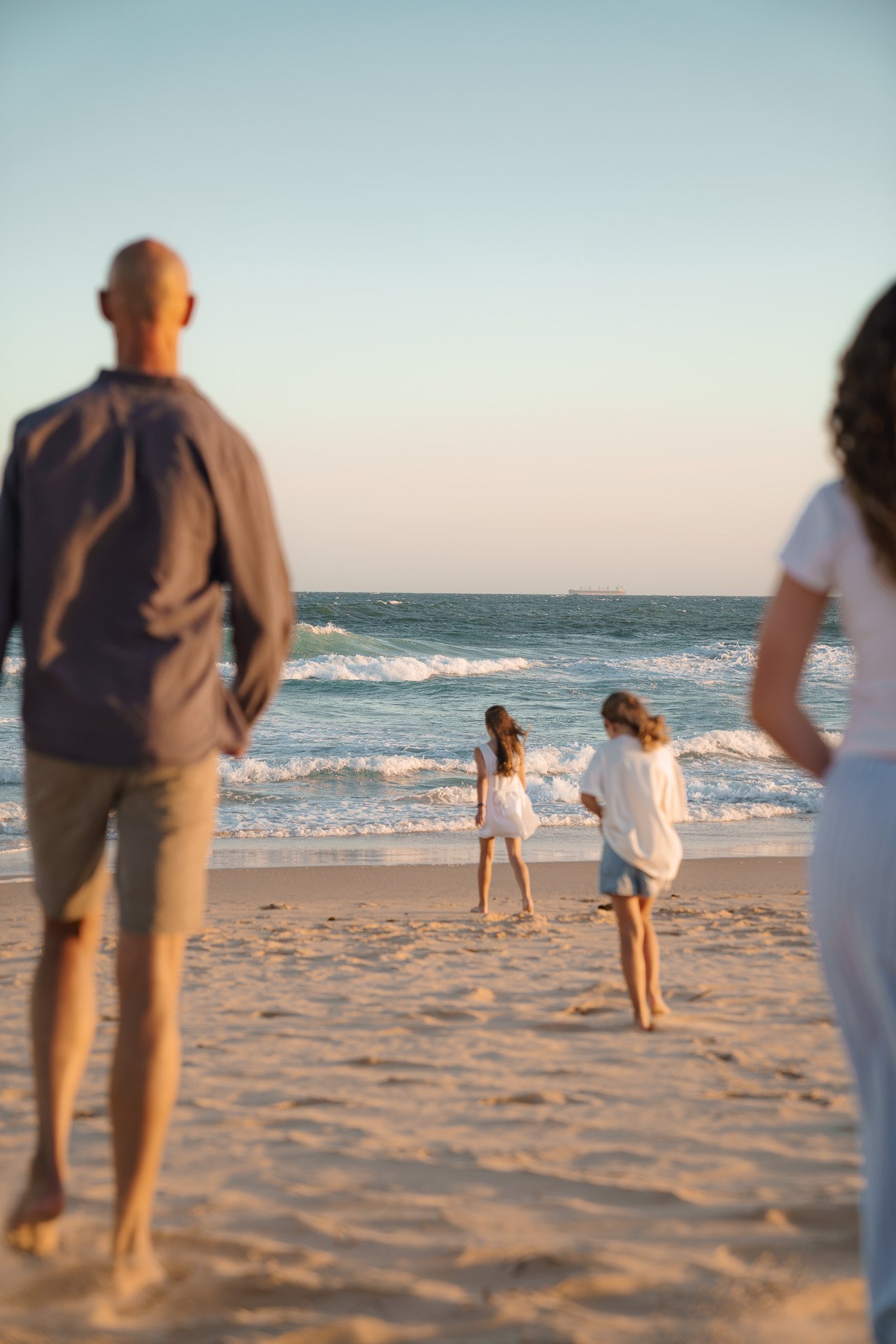 People on a sandy beach with two young girls near the water, and two adults in the foreground, watching the scene.