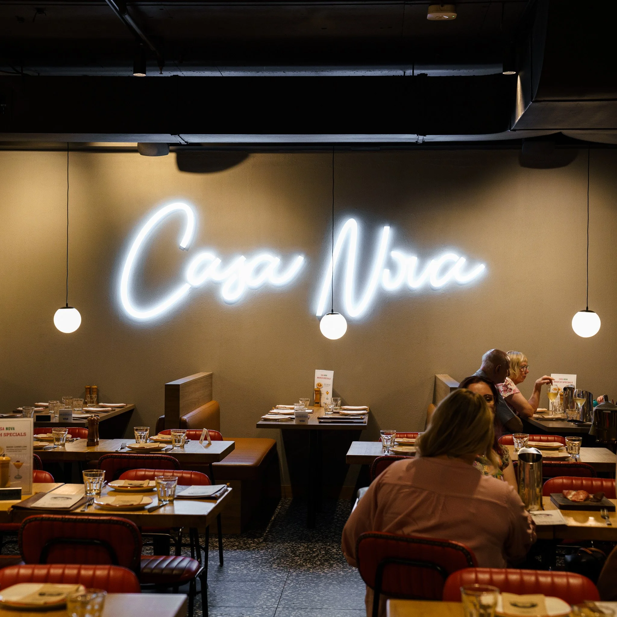 Inside a restaurant with a neon sign on the wall that reads 'Casa Nova.' There are tables set with plates, glasses, and napkins, with some patrons dining and talking.