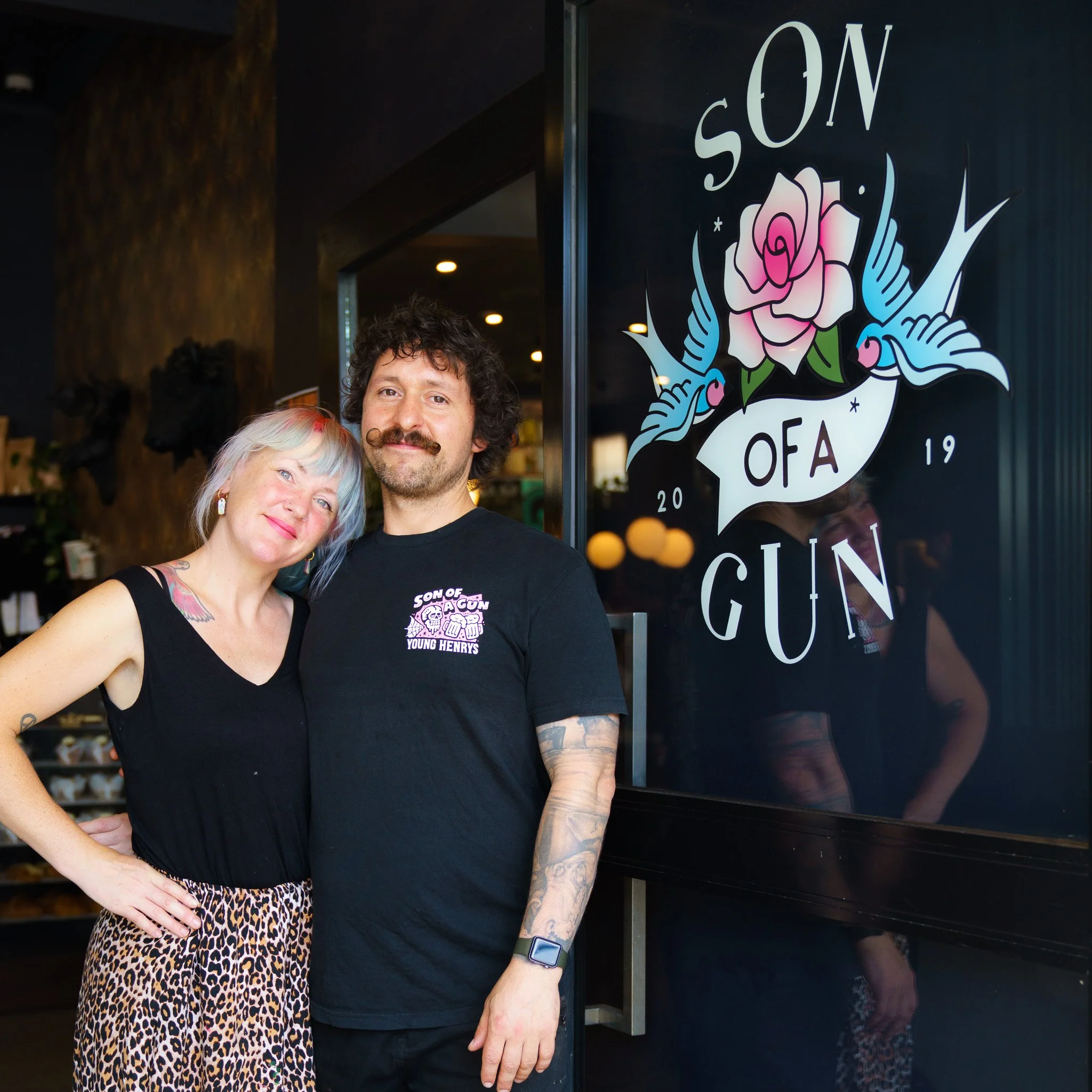 A man and woman standing together outside a store with a sign that reads "Son of a Gun" and features a pink rose with blue leaves. The woman has short, multi-colored hair and is wearing a black sleeveless top and leopard print pants. The man has curly hair, a mustache, tattoos on his arms, and is wearing a black T-shirt with a logo that also says "Son of a Gun".