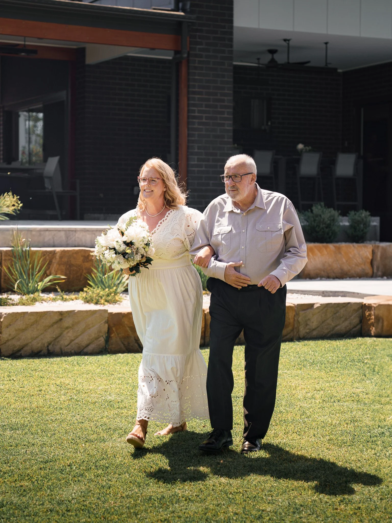 A woman in a white dress holding a bouquet is walking arm-in-arm with an older man in a light shirt and dark pants on a grassy lawn outside a modern house.