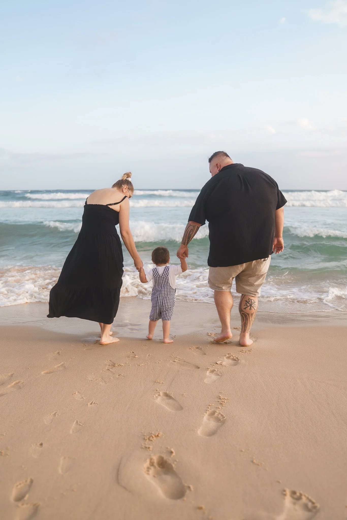 A family of three walking hand in hand on the beach near the ocean waves, with footprints in the sand.