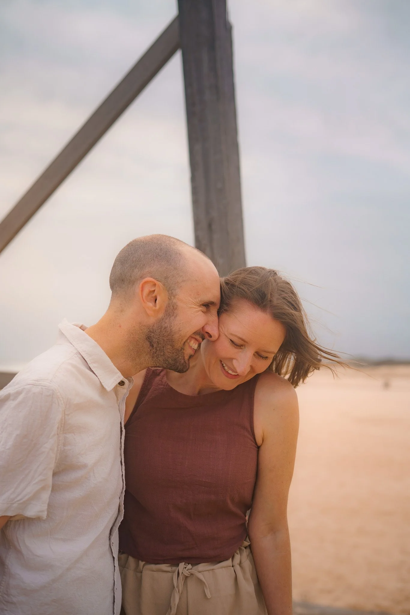 A smiling man and woman leaning close together outdoors, with a large wooden cross in the background and a cloudy sky.