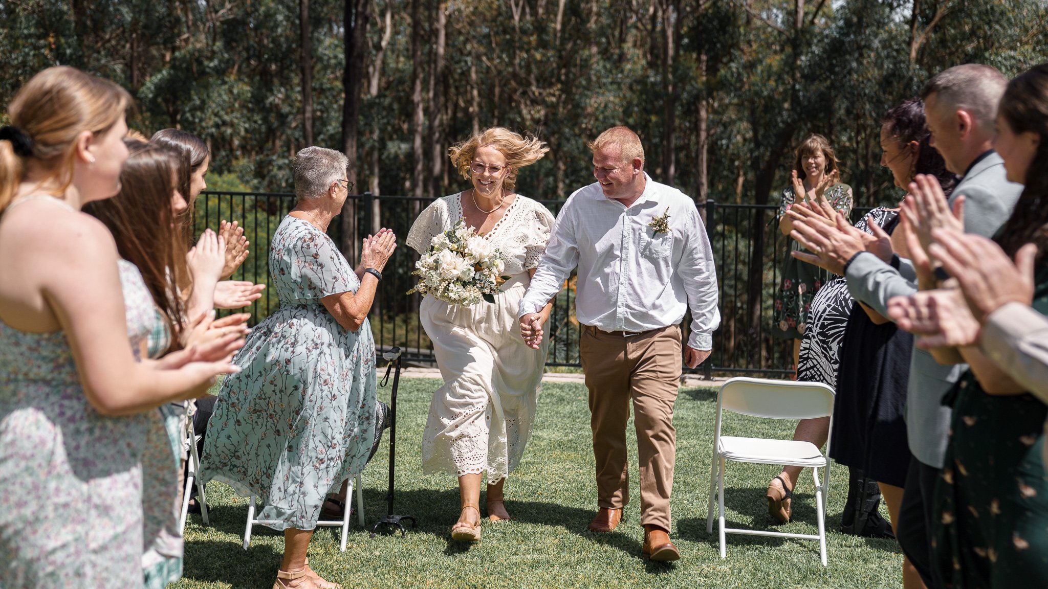 Couple walking down aisle holding hands, surrounded by clapping guests at outdoor wedding ceremony in a green, wooded area.