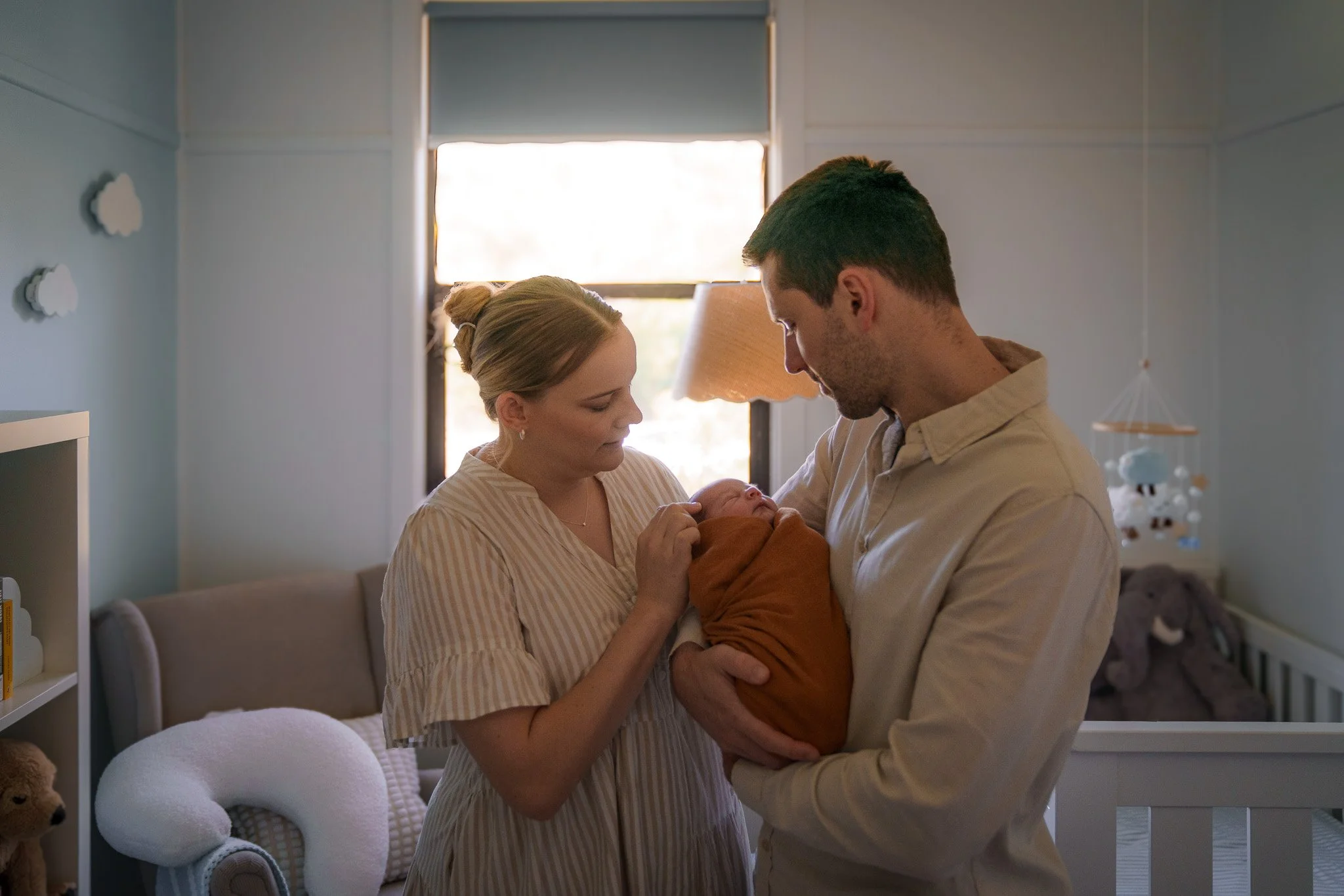 A family stands in a nursery holding a newborn baby. The mother and father look at the baby lovingly, with warm lighting coming through the window in the background.