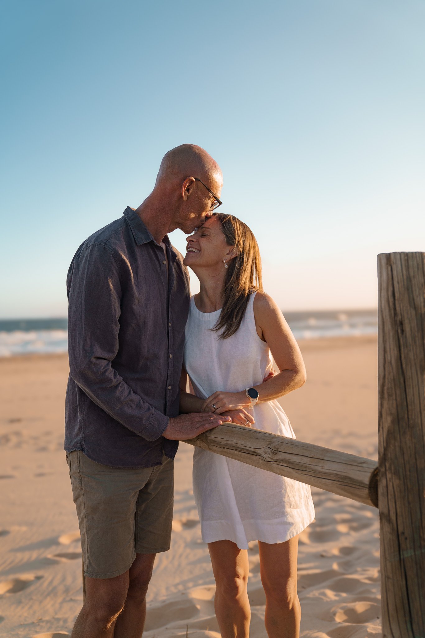 A couple standing on a beach during sunset, embracing and smiling at each other.