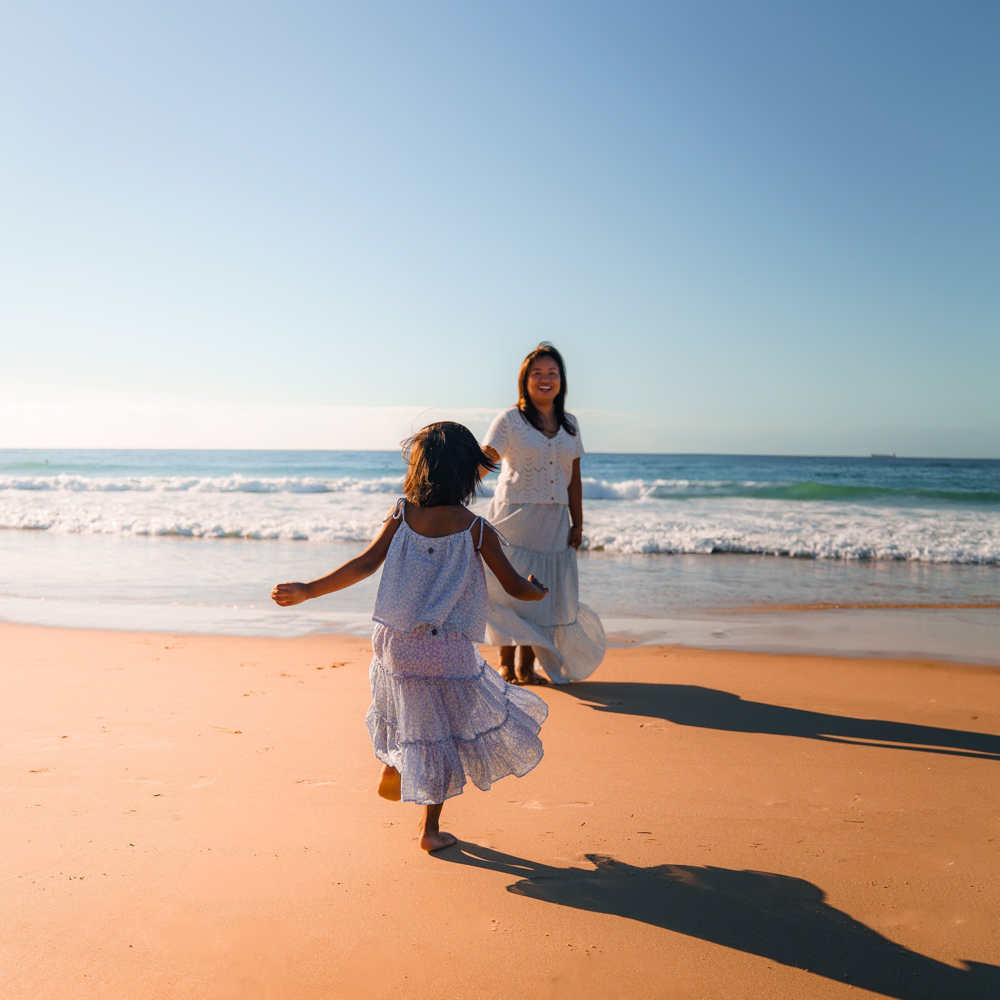 A woman and a young girl playing on a sandy beach during sunset, with ocean waves and a clear sky in the background.