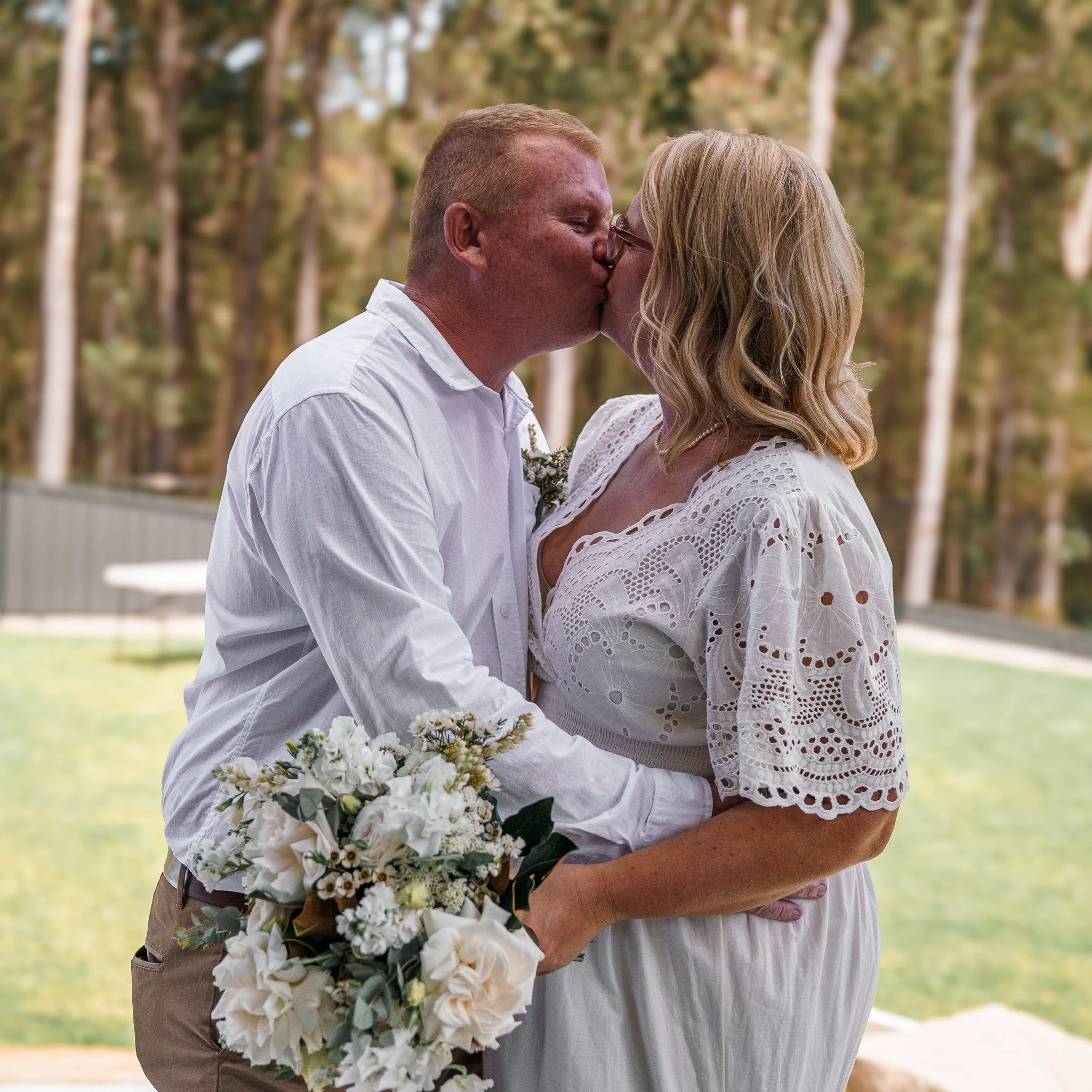 A couple sharing a kiss outdoors during a wedding or special occasion, with the woman holding a bouquet of white flowers, and both dressed in white attire, surrounded by trees.