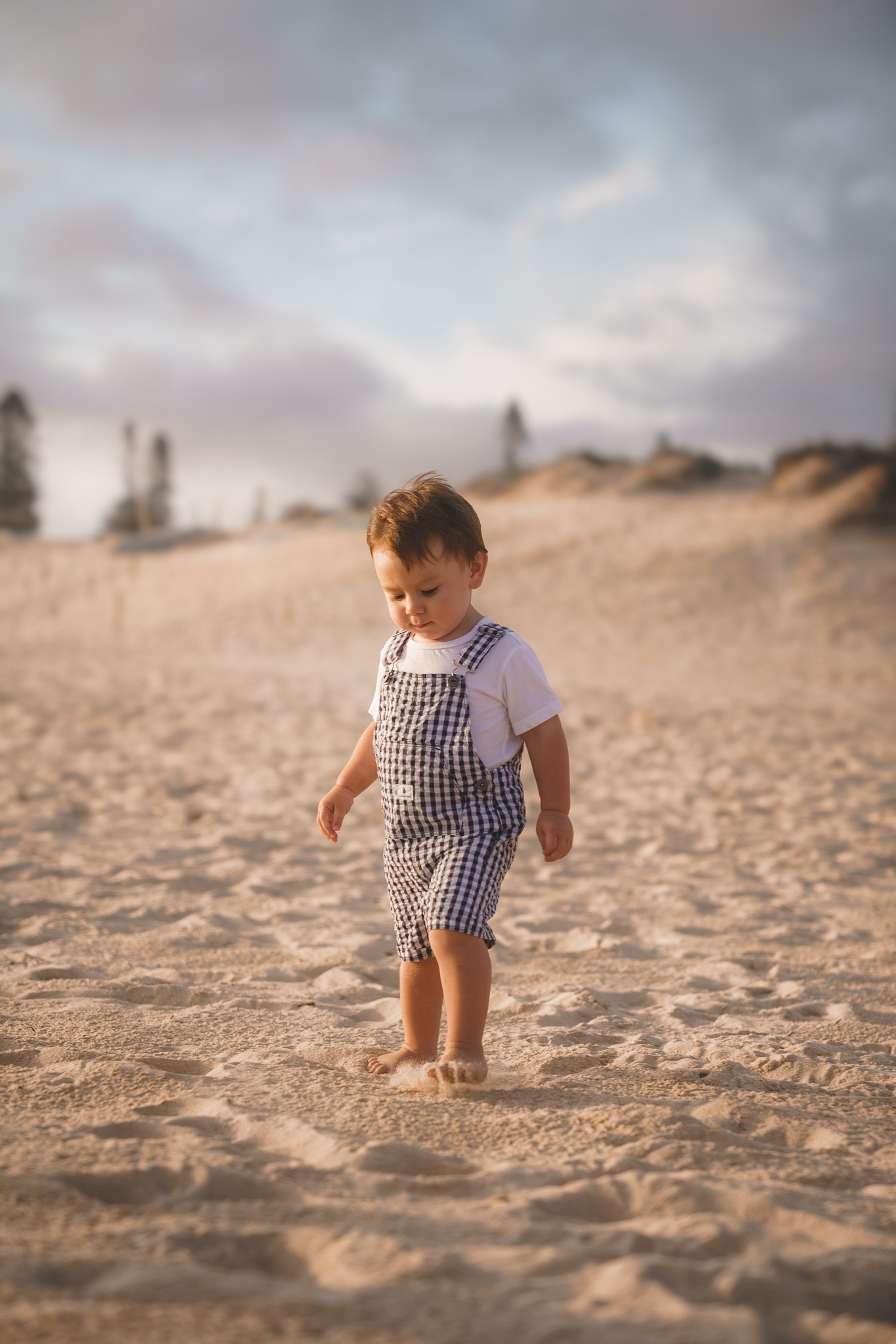 Young boy walking barefoot on sandy beach with dunes and cloudy sky in the background.