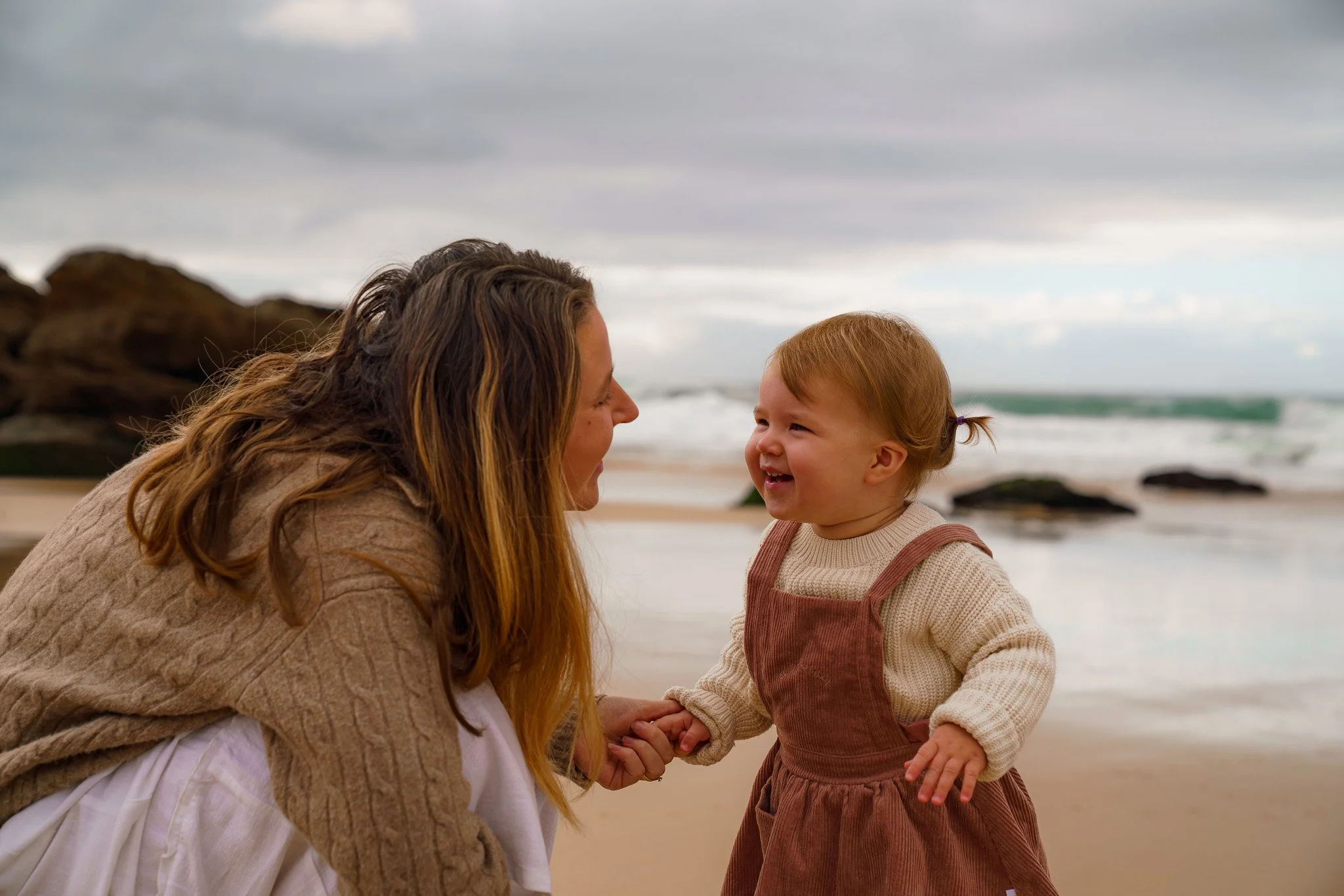 A woman and a young girl holding hands on a beach with rocks and ocean in the background, both smiling at each other.
