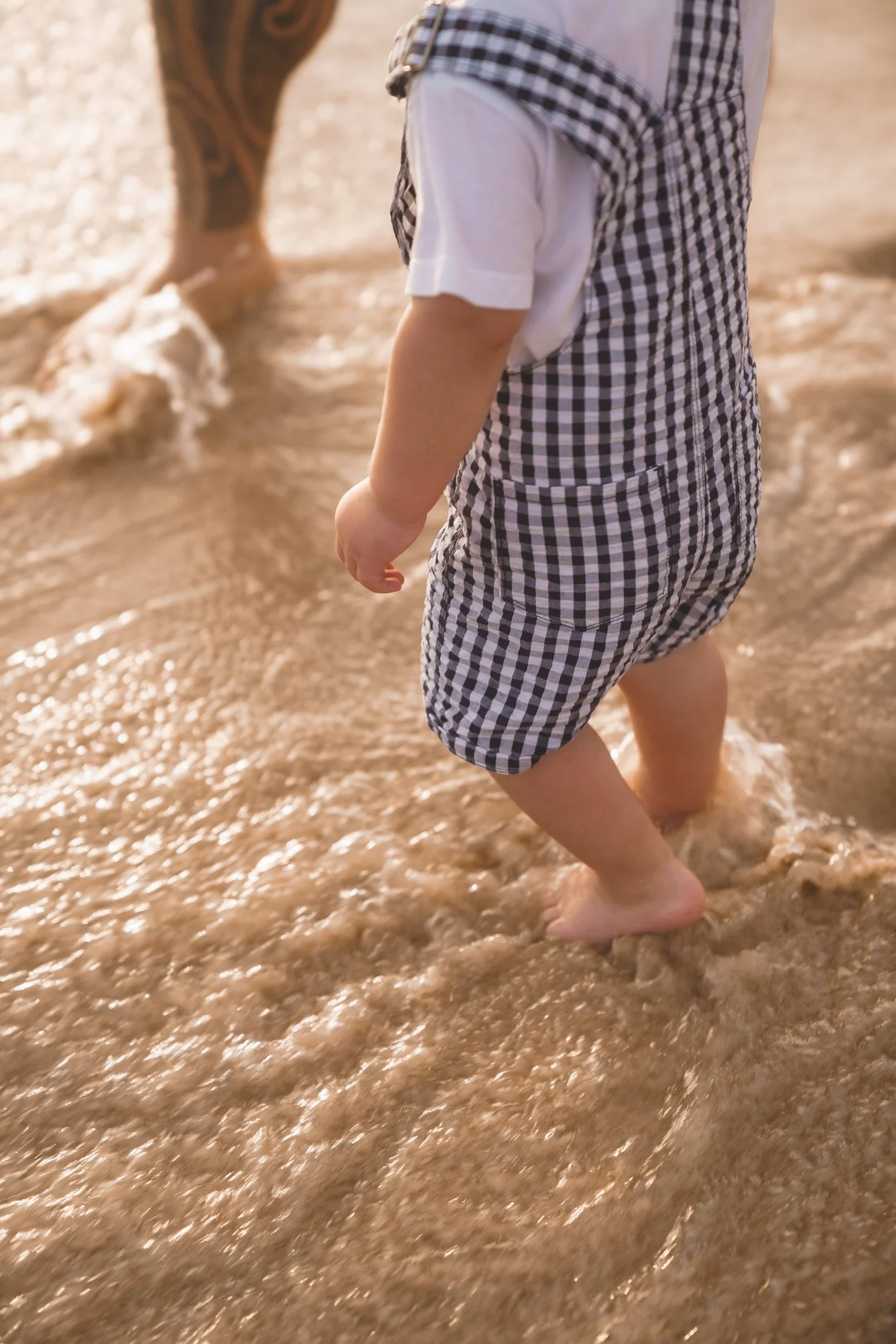 A young child with short hair, wearing a checkered vest and white shirt, wading barefoot in the shallow water of a beach.