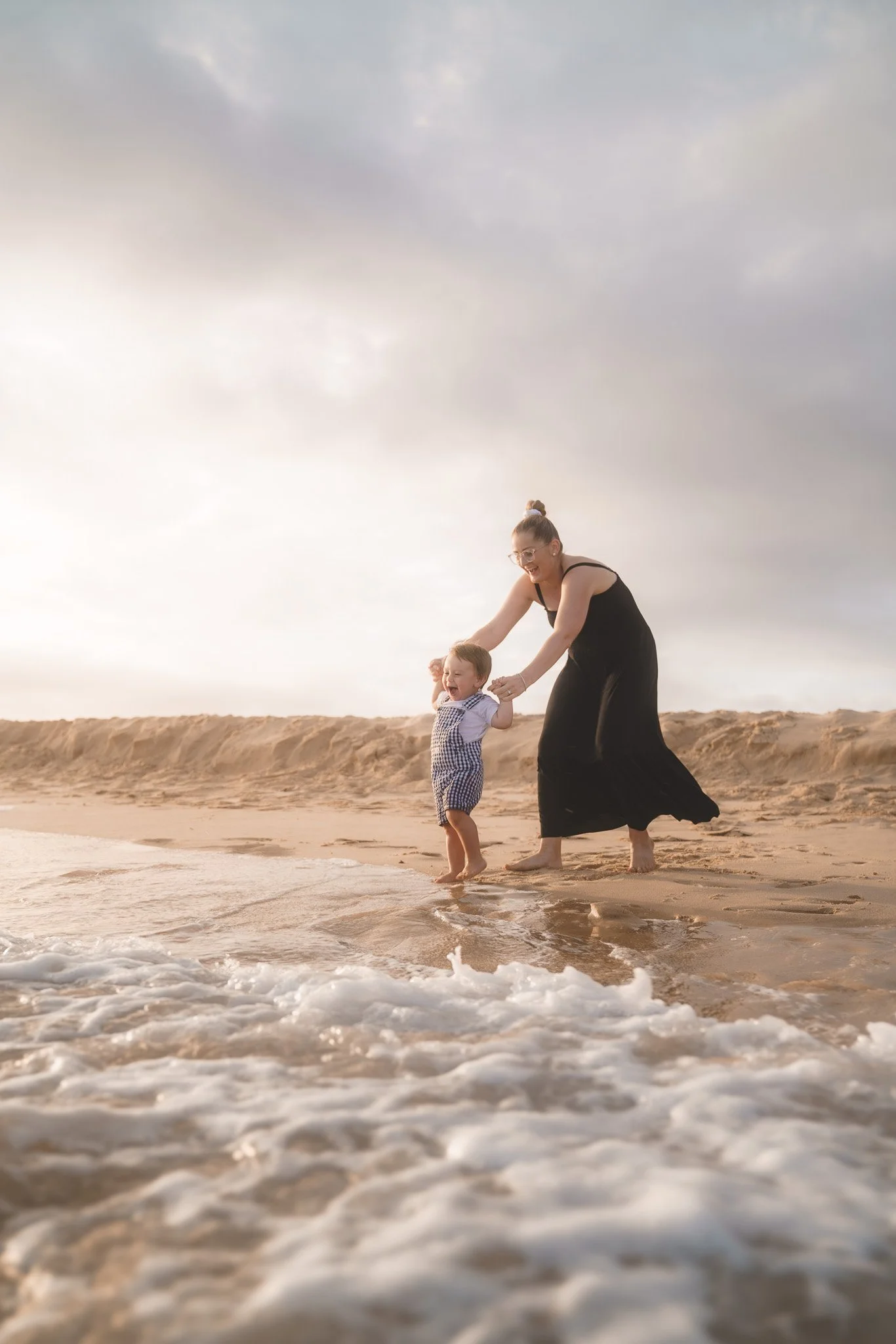 A woman and a young boy playing at the beach, holding hands and smiling, with the shoreline and cloudy sky in the background.