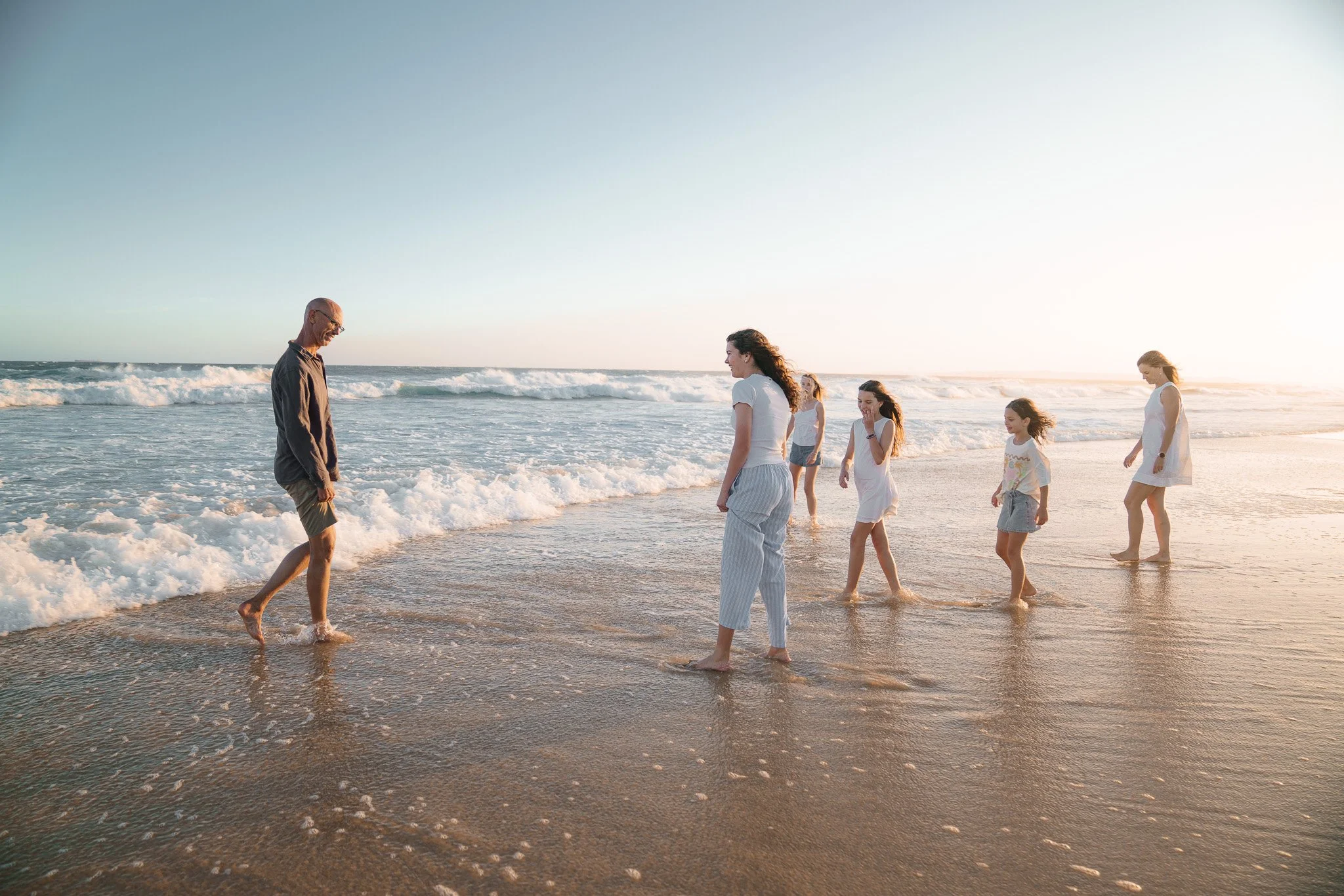 A group of six people, including children, walking along the shoreline at the beach during sunset, with gentle waves and a clear sky.
